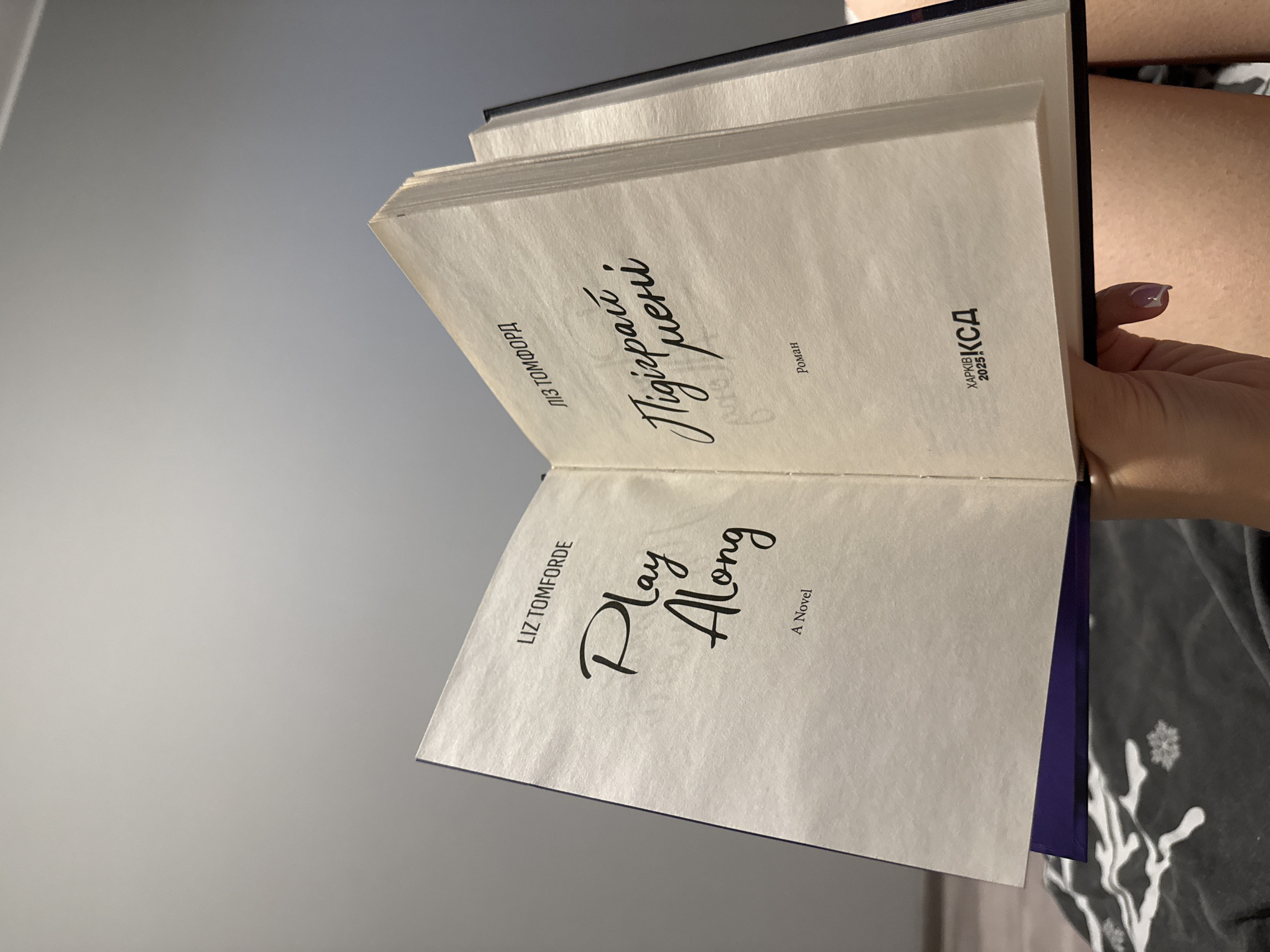 An open book is held by a person's hand, revealing two pages. The left page displays "LIZ TOMFORDE" at the top, followed by the title "Play Along" in a stylized script, and "A Novel" below. The right page features "ЛІЗ ТОМФОРД" at the top, then the title "Підіграй мені" in a similar script, and "Роман" below. At the bottom of the right page are the words "ХАРКІВ" and "2025" next to a logo that reads "КСД". The book's cover appears to be dark blue or black. The background is a plain, light gray wall. The person's hand has a fingernail with a purple-ish polish. The image was taken indoors, in what appears to be a domestic setting, with no identifiable landmarks or features to indicate its location within the city of Łódź, Poland.