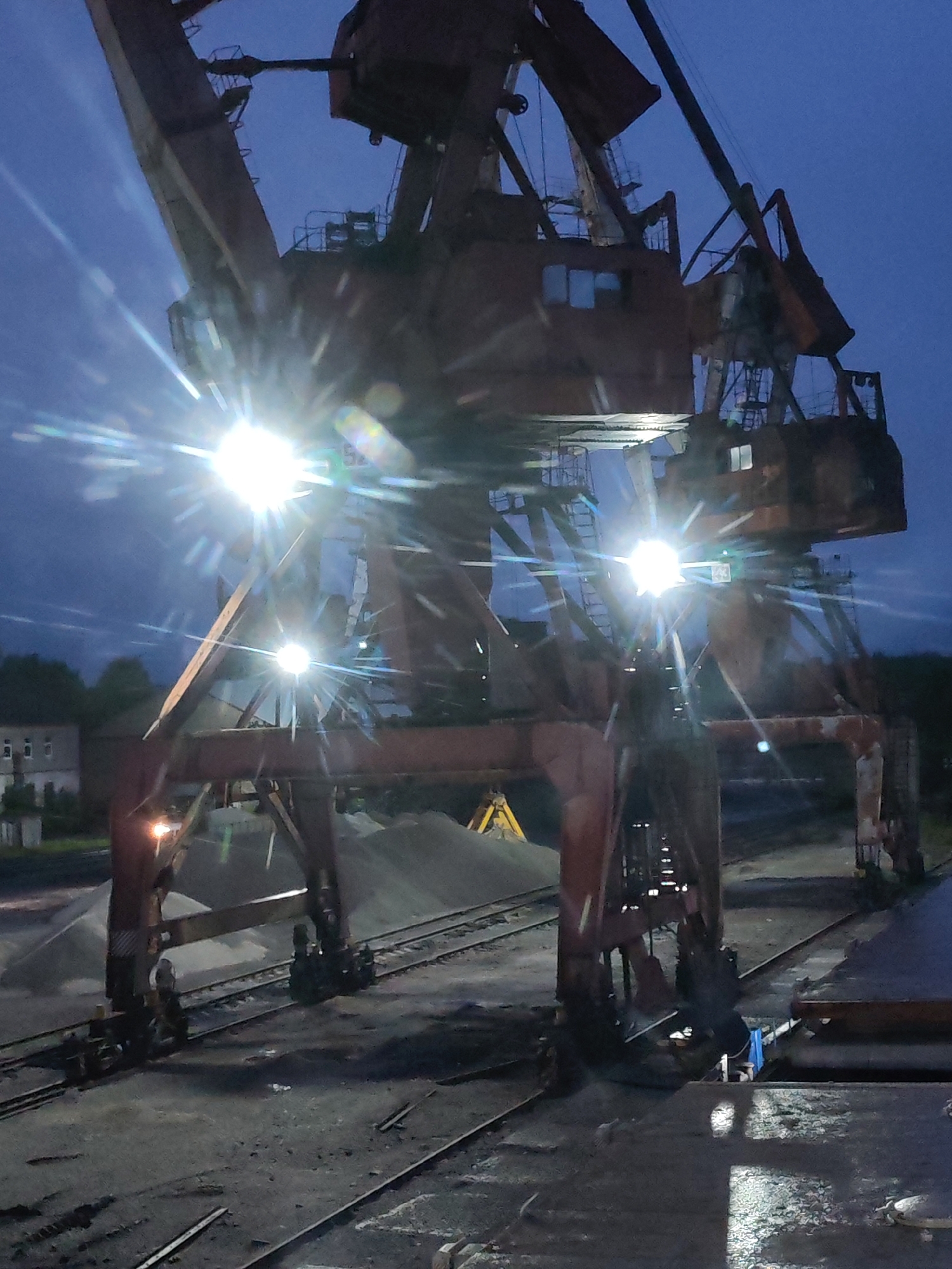 The image depicts a large, weathered industrial gantry crane at night or late dusk/early dawn in what appears to be an industrial port or railway yard in Medvezhyegorsk, Russia. The dominant subject is the crane, a substantial structure made of rusty, dark-colored steel beams, featuring multiple support legs, cross-braces, and operator cabins.
Bright artificial lights are mounted on various parts of the crane, casting intense illumination and producing prominent star-shaped lens flares that pierce the dark blue sky. The sky itself shows faint cloud patterns. Below the crane, several parallel railway tracks are visible on a dark, possibly wet, ground surface that reflects some of the artificial light.
In the background, behind the crane's supports, are large piles of light-colored aggregate, such as gravel or sand. A piece of yellow heavy machinery, possibly an excavator or loader, is partially visible behind one of these piles. To the far left, a multi-story building with several windows can be seen against the horizon, partially obscured by trees or foliage.
No direct human activity is apparent, suggesting the crane is stationary or awaiting operation. The scene conveys a stark, industrial atmosphere. The rusty appearance of the crane indicates its age and consistent use in a demanding environment. No discernible text is visible on any objects within the photograph.