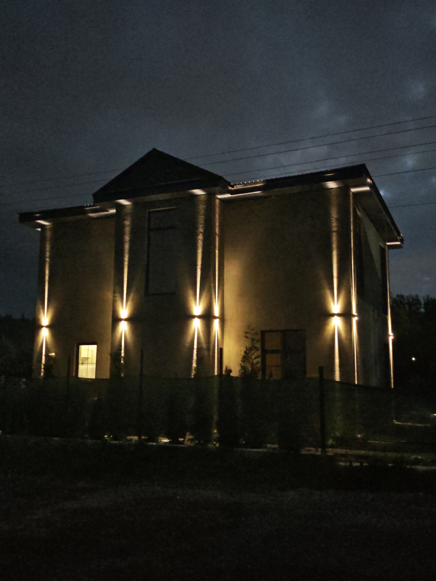 The image depicts a modern, two-story building with a dark, overcast sky. The building is illuminated by vertical, upward-facing wall lights that cast strong beams of light onto its beige exterior. There are several windows visible, with one on the lower left side glowing from within. A dark fence runs along the front of the property, with some dark green shrubbery behind it. The ground in front of the fence is dark and appears to be grass. In the background, dark trees are visible. Overhead, power lines are strung across the sky. The overall mood is somewhat dramatic due to the strong lighting and dark, cloudy sky. It appears to be nighttime. The location is identified as Надежда Снт, Russia. There is no text visible in the image.