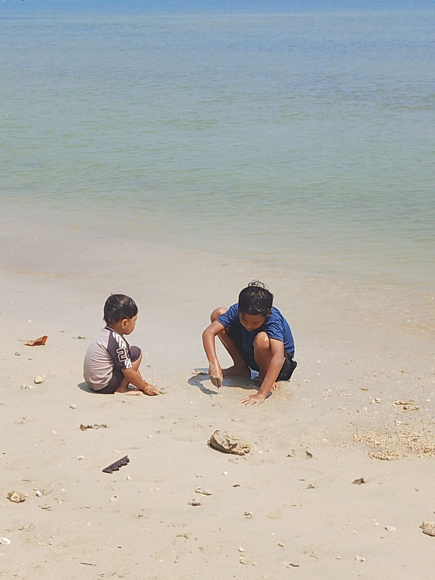 Two young boys are playing on a sandy beach in Pejagan, Indonesia. The boy on the left is crouched down, facing to the right. He is wearing a grey and black t-shirt with white accents and dark shorts. The boy on the right is also crouched down, facing forward and slightly to the left. He is wearing a blue t-shirt and black shorts, and his hands are in the sand. The background consists of calm, shallow water with a clear blue sky above. The sand is light brown and scattered with small shells and debris.