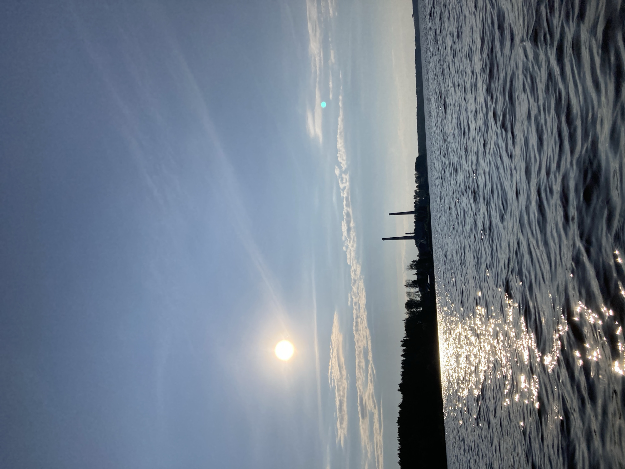 The image captures a serene natural landscape. A bright sun, casting a warm glow, is visible in the clear sky, partially obscured by wispy clouds. The sun's reflection shimmers on the rippling surface of a large body of water, creating a dazzling effect. The water extends to the right of the frame, while the left side shows a distant treeline silhouetted against the sky. Two tall, industrial smokestacks stand prominently against the horizon, suggesting the presence of a factory or power plant. The scene evokes a sense of tranquility and the beauty of nature, possibly at sunset or sunrise due to the low angle of the sun and the soft lighting. The time of day appears to be late afternoon or early evening. The location is identified as Chaikovsky, Russia, which aligns with the visual cues of industrial elements and a natural setting. There is no visible text in the image.