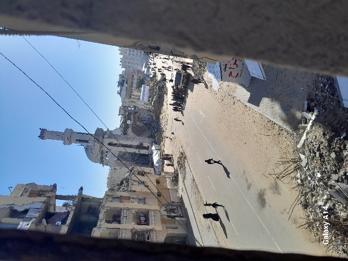 The image displays an extensively damaged urban streetscape in Khan Yunis, Palestinian Territory, under a clear blue sky. A prominent white mosque with a tall minaret, partially destroyed and missing sections of its dome and structure, stands in the background. Adjacent and opposite the mosque, numerous multi-story buildings exhibit severe damage, with facades partially collapsed, exposed interiors, and significant rubble accumulation at their bases.
A paved two-lane street, partially covered with sand and debris, traverses the scene. Multiple individuals, including adults and children, are walking on the street. In the foreground's left lane, three figures are visible, one carrying a light-colored object. Further down the street, larger groups of people are congregated, particularly on the right side where a white pickup truck is parked near one gathering. Makeshift tarpaulin shelters are discernible amidst the debris near the mosque. Sunlight casts distinct shadows across the street and structures, indicating daytime. The ground in the immediate foreground is covered in rubble and broken building materials.