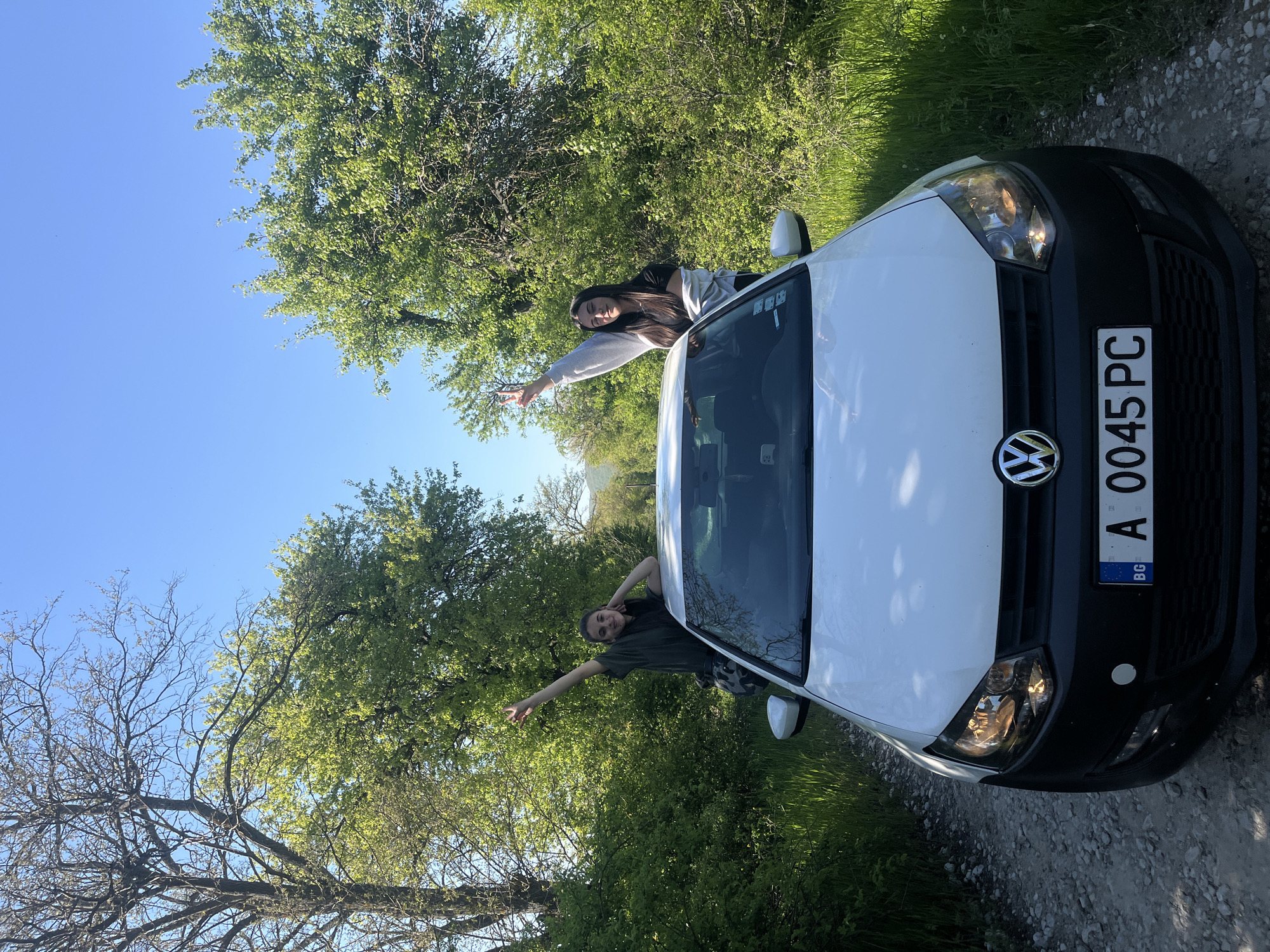 The image shows a white Volkswagen car parked on a road adjacent to a lush, green wooded area under a clear, blue sky. Two individuals are interacting with the car, each leaning out of a window and gesturing with their hands. The car's license plate is visible, bearing the Bulgarian flag and the alphanumeric identifier "A 0045 PC". The scene suggests an outdoor location in or near a wooded area, with the clear sky indicating a sunny day. The road appears to be a relatively quiet, unpaved surface. The city of Rish, Bulgaria, is not discernible from the image alone, although the Bulgarian license plate indicates that the location is within Bulgaria.