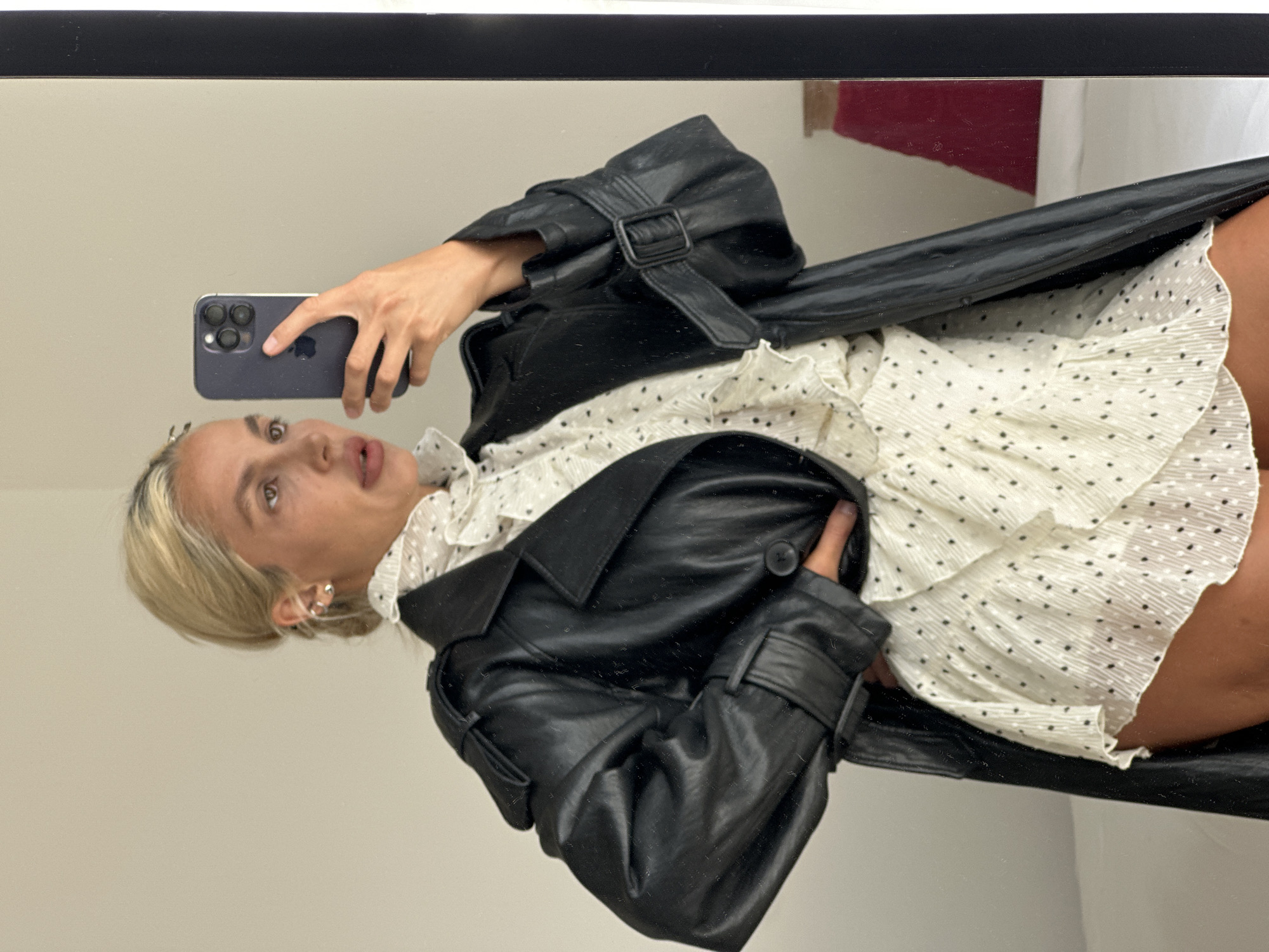 A woman is reflected in a mirror, holding a phone to take a selfie. She is wearing a white ruffled blouse with a polka dot pattern and a black leather trench coat. Her hair is pulled back in a bun. The background is a plain wall, and a bed is visible in the reflection. The location is Kuta Selatan, Indonesia.
