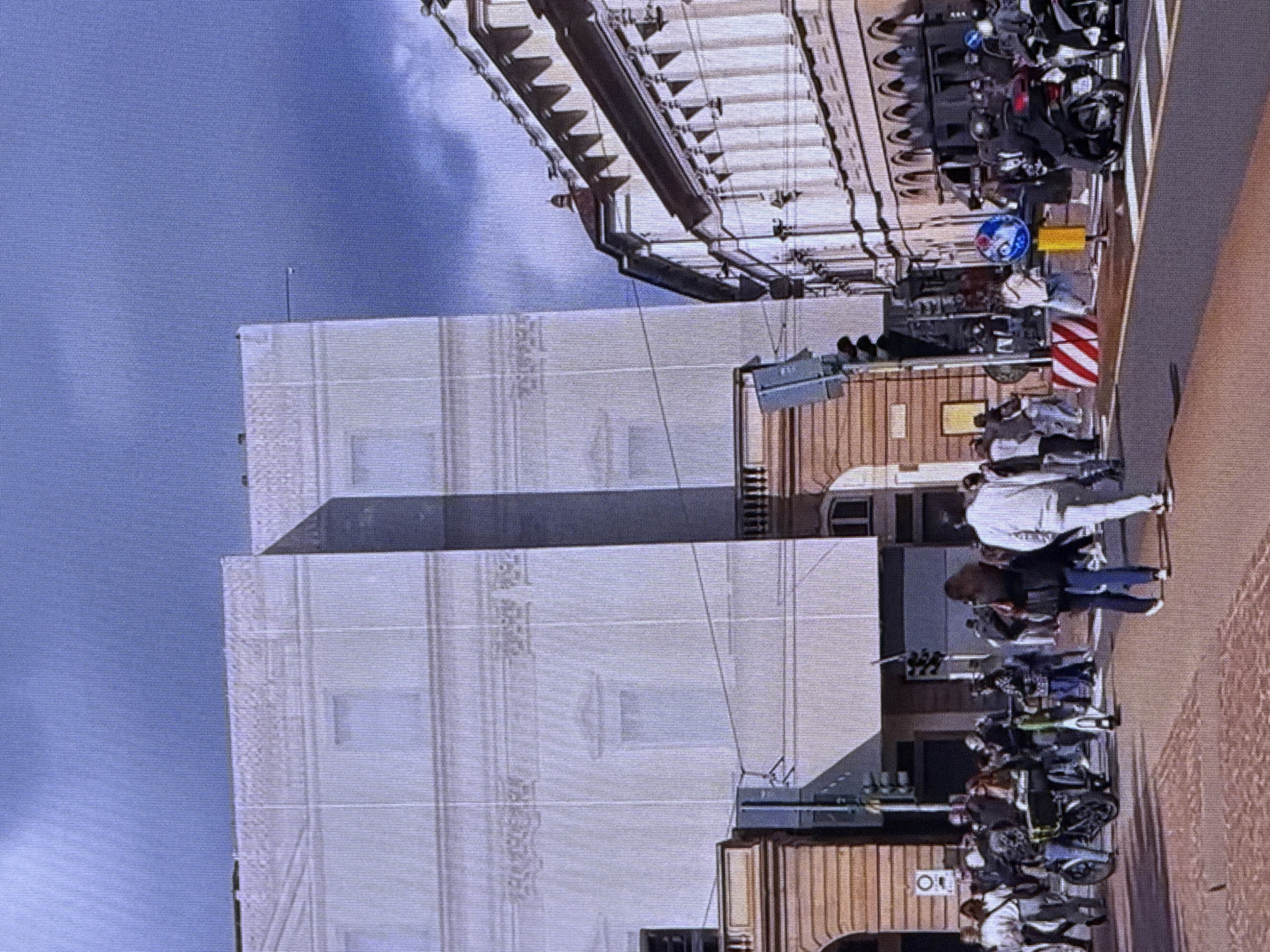 The image depicts a street scene with several people and vehicles in front of buildings. The main building on the left is partially covered by a white tarp, suggesting construction or renovation. A street runs through the foreground with a pedestrian crossing.
On the left side of the street, a person is walking alongside an electric scooter. Further back, a group of people are visible on the sidewalk. In the center of the image, a man in a white tracksuit is walking across the street, and other pedestrians are also present. To the right, several people are on motorcycles or scooters, waiting at what appears to be an intersection.
The architecture of the buildings suggests an urban environment. The building on the right has classical architectural elements with columns and arched windows. The sky is partly cloudy with a mix of blue and grey. The overall impression is that of a busy city street during the daytime.
This scene does not resemble the city of Dubai, United Arab Emirates. The architecture, streetscape, and general ambiance are not characteristic of Dubai.