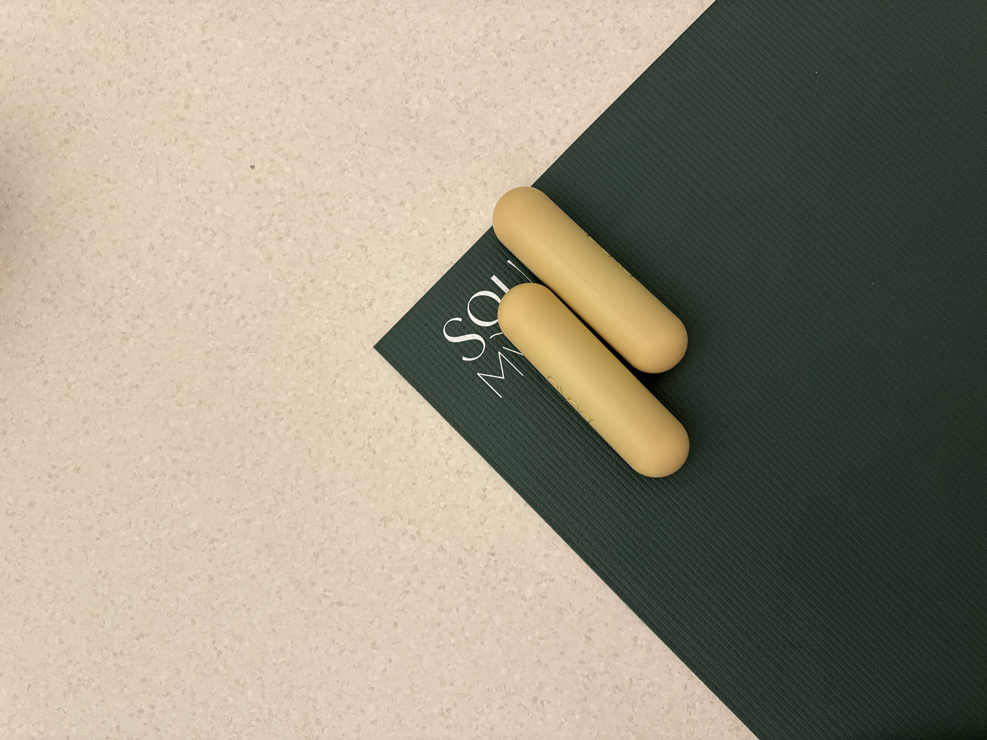 The image depicts two light yellow suppositories lying on the corner of a dark green, ribbed yoga mat. The yoga mat is positioned on a light beige countertop with a speckled surface. The text "SOL" is visible on the yoga mat, suggesting the brand name of the mat. The lighting appears to be artificial and even. The scene does not provide any specific indicators of being located in Dubai, United Arab Emirates.