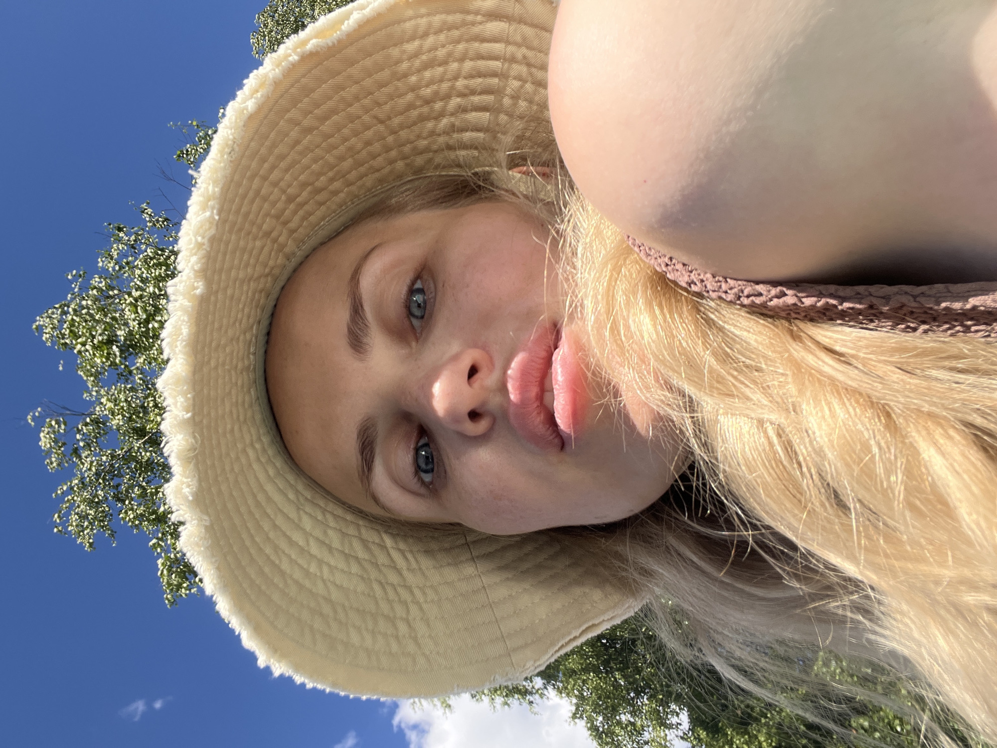 A young woman with long, blonde hair and blue eyes is captured in a selfie outdoors. She is wearing a wide-brimmed, straw-colored hat with a frayed edge. A portion of her chest is visible, clad in a textured, pinkish-brown garment, possibly a bikini top or dress. Her lips are slightly parted, and her gaze is directed towards the camera. The background features a bright blue sky with some scattered white clouds, and the lush green foliage of trees is visible, suggesting a warm, sunny day. The perspective of the photo, with the sky and trees appearing above the woman's head, indicates she might be lying down or looking up. The scene evokes a sense of leisure and outdoor enjoyment, perhaps at a park or beach. The lighting suggests it is daytime, with the sun casting a warm glow. There is no visible text in the image.