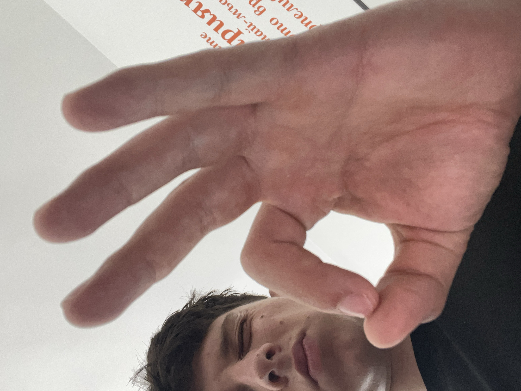 The image is an extreme low-angle shot, looking upwards at a person and their hand. In the foreground, a right hand is prominently displayed, making a gesture where the thumb and forefinger form a circle, while the other three fingers are extended. This close-up perspective causes the hand to appear large and dominant in the frame.
Below the hand, in the lower section of the image, the upper part of a person's face is visible, including dark hair, part of their forehead, eye, and nose. Their gaze seems directed upwards towards the camera. The person is wearing a dark, likely black, piece of clothing, visible on the right side of the frame.
The background consists of a bright, plain white surface, presumably a ceiling or wall, suggesting an indoor setting. Blurred text in a red-orange color is partially visible on this white surface. The script appears to be Cyrillic, with discernible fragments such as "me", "puru" (resembling "пури"), "най-мъ", "бъ", "ou", and "neay". These textual clues are consistent with the provided location context of Zlatograd, Bulgaria. The lighting is bright and even, indicating a well-lit environment. The overall impression is a casual, possibly self-taken photograph featuring a playful hand gesture.