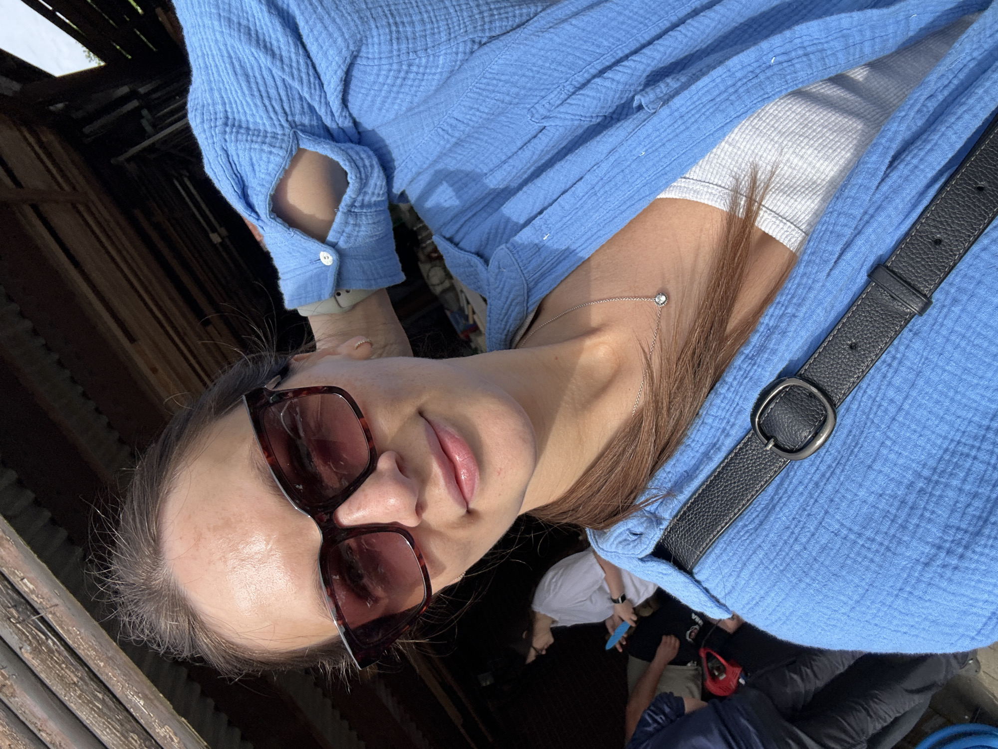 A woman is posing for a selfie. She is wearing sunglasses and a blue shirt over a white top. She also has a black bag slung across her body. The background is a dimly lit, possibly enclosed space with wood paneling. Other people are visible, but they are out of focus. The location is Bargstedt, Germany.