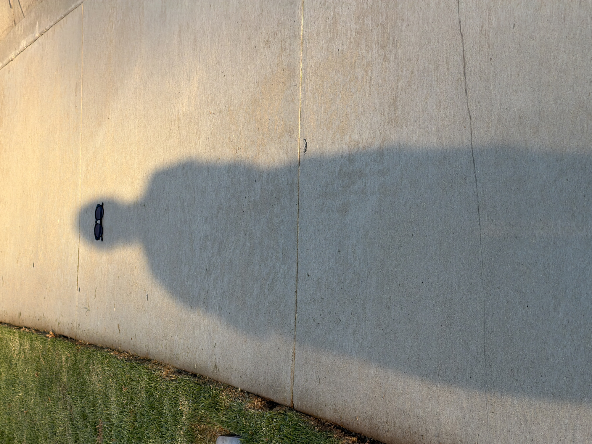The image shows a long shadow of a person on a concrete sidewalk, with a pair of sunglasses resting on the sidewalk within the shadow. To the left of the sidewalk, a strip of green grass is visible. The scene is illuminated by warm, directional light, suggesting it was taken during the golden hour. The concrete appears to be a typical urban or suburban sidewalk. The location is Aliso Viejo, United States of America.