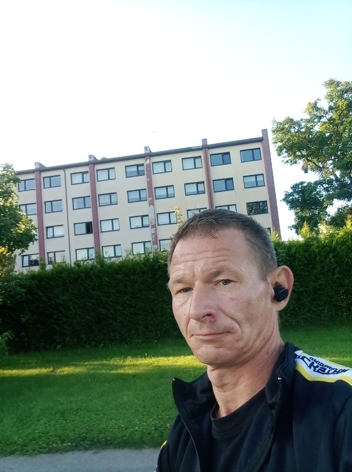 In Tapa, Estonia, a mid-shot selfie captures a man with short, graying hair and fair skin. He has a prominent scar on his nose and appears to be wearing a black t-shirt under a black zip-up jacket with yellow and white detailing on the shoulders. A black wireless earbud is visible in his right ear.
The man is positioned in the foreground, with a slightly out-of-focus view. Behind him, a multi-story building with a light-colored facade and numerous rectangular windows is visible. The building appears to be residential or office complex, with several floors. A dense, dark green hedge forms a barrier across the middle ground of the image.
To the left of the building, the top of a leafy green tree is visible, and to the right, another mature tree with lush green foliage stands. The ground in the foreground consists of a paved pathway or road on the bottom, transitioning to a well-maintained grassy area with dappled sunlight. The sky is bright and clear, suggesting daytime. The overall scene appears to be an outdoor urban or suburban environment.