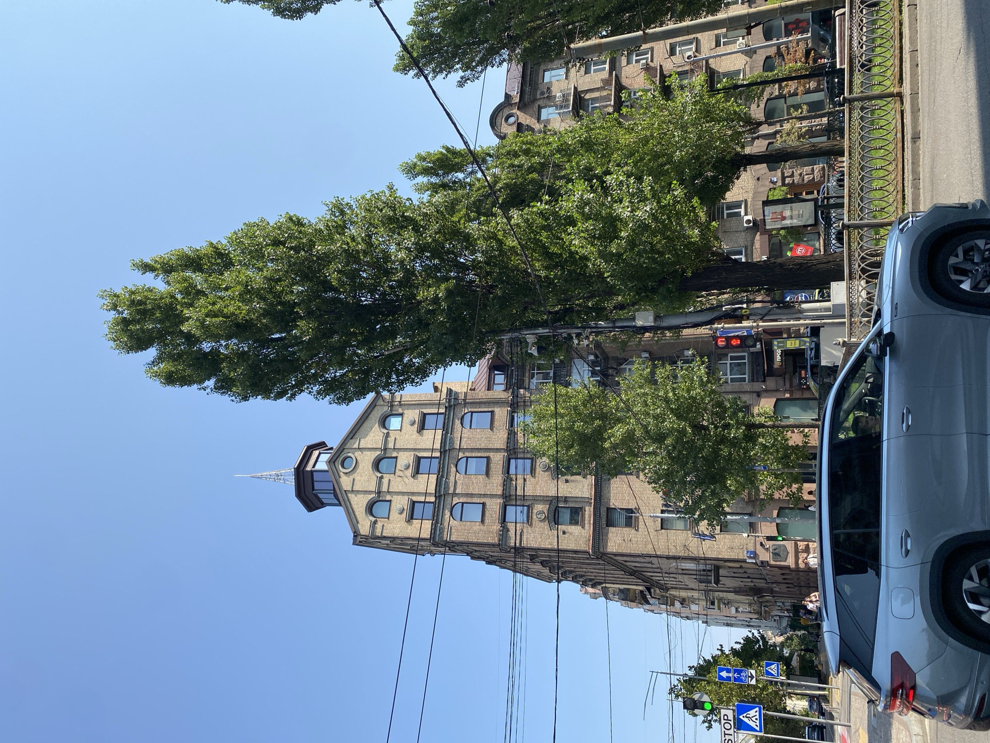 A medium-shot, eye-level, outdoor photograph shows a street scene with a multi-story brick building and trees under a clear blue sky. The building, with a pointed spire on top, is made of light brown brick and has many windows, some arched and some rectangular. Tall, slender trees with lush green leaves flank the building. In the foreground, a silver SUV is driving on the street, and a pedestrian crossing with traffic lights and signs is visible to the left. Overhead power lines crisscross the sky. The overall impression is of a sunny day in an urban environment.