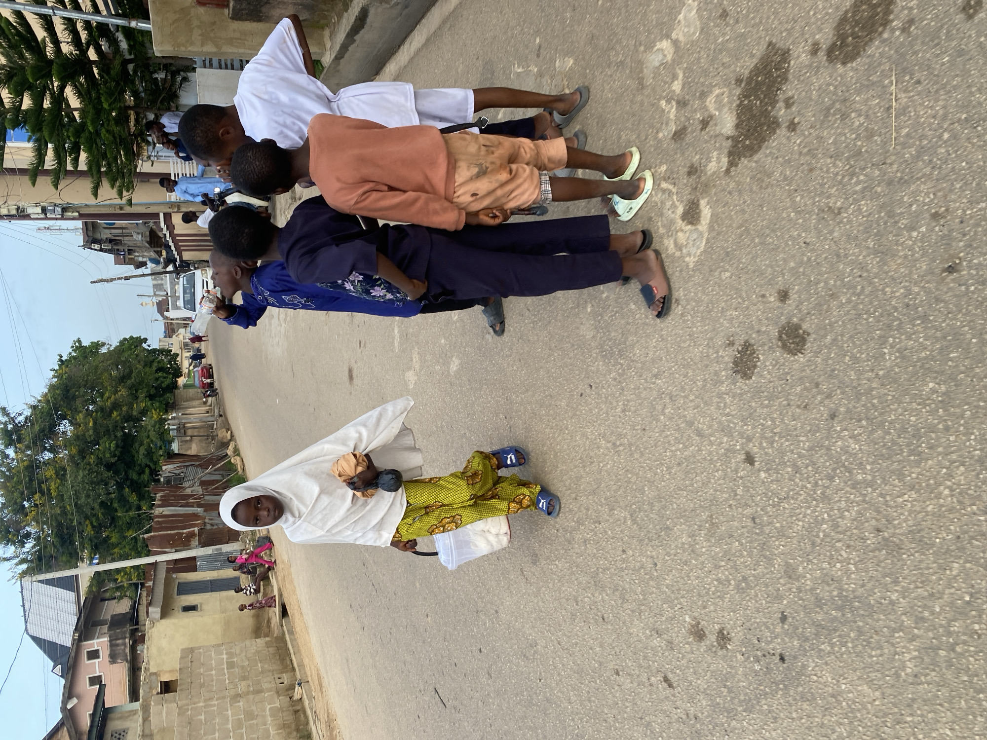 The image depicts a street scene in Jos, Nigeria, with several children present. In the foreground, a young girl wearing a white hijab and patterned yellow trousers walks towards the camera, carrying a plastic bucket in her left hand and a bundle wrapped in cloth in her right. She is wearing blue slide sandals.
Behind her, a group of boys stands on the asphalt road. One boy in a blue traditional outfit is drinking from a plastic bottle. Another boy, wearing a dark blue long-sleeved shirt and dark trousers, stands beside him. Further to the right, a boy in an orange long-sleeved shirt and light-colored shorts is visible, and another boy in a white long-sleeved shirt and white shorts stands next to him. The boys are wearing various types of footwear, including sandals and slides.
In the background, buildings line the street, some appearing to be residential with corrugated iron roofs and brick or stucco walls. Trees and vegetation are also visible, contributing to the urban landscape. A red car is parked further down the street. The overall lighting suggests it is daytime, possibly late afternoon. The scene captures a moment of everyday life on a street in Jos.