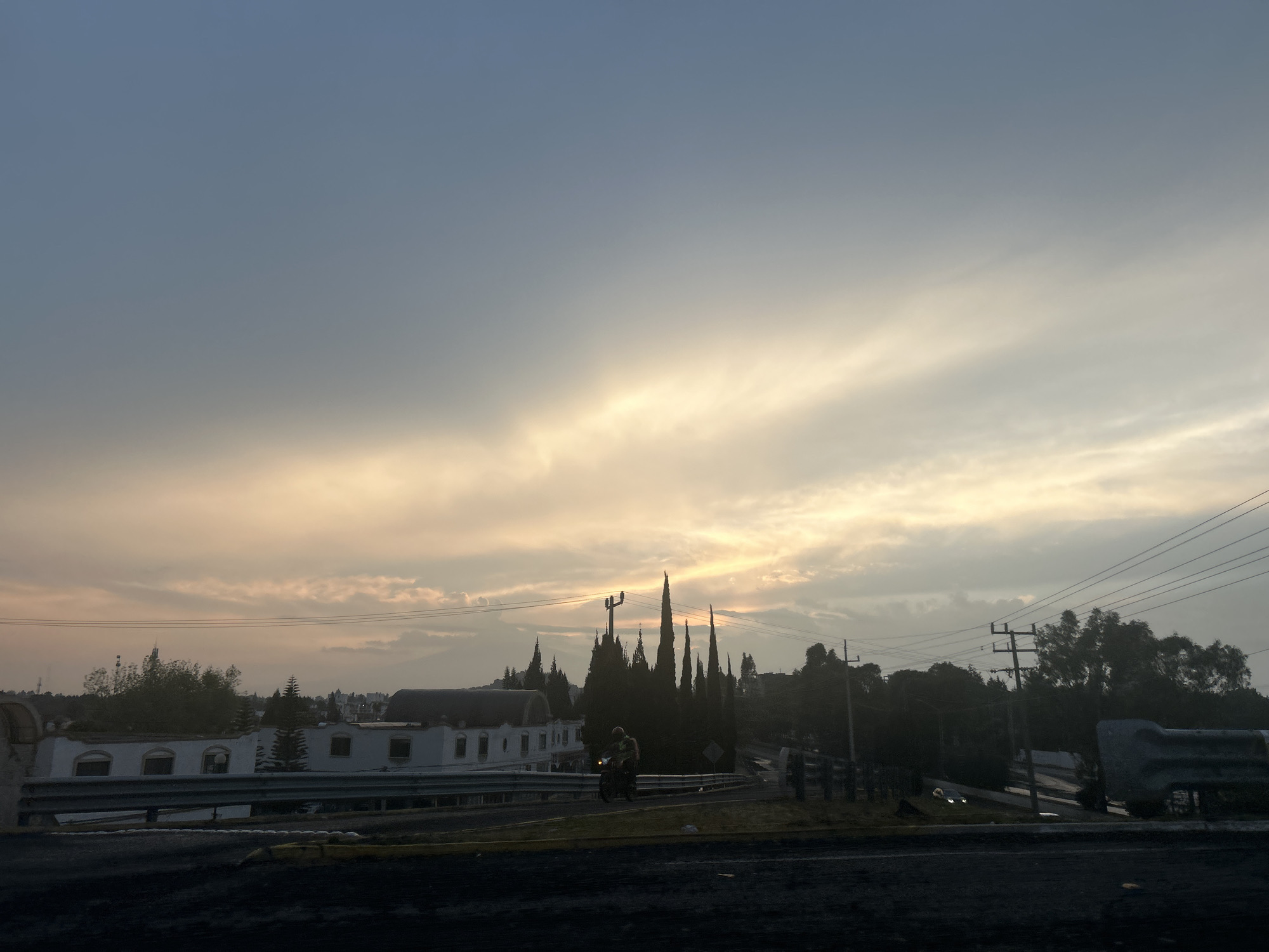 The image depicts a silhouetted cityscape at dusk or dawn under a sky with a mix of blue and golden clouds. A two-story white building with multiple windows is visible on the left, protected by a roadside barrier. Tall, slender trees stand prominently in the center, partially obscuring a utility pole with power lines stretching across the sky. A lone motorcyclist is present on the road, traveling away from the viewer. To the right, a cluster of trees and another utility pole are seen. The foreground is dominated by the dark surface of the road.