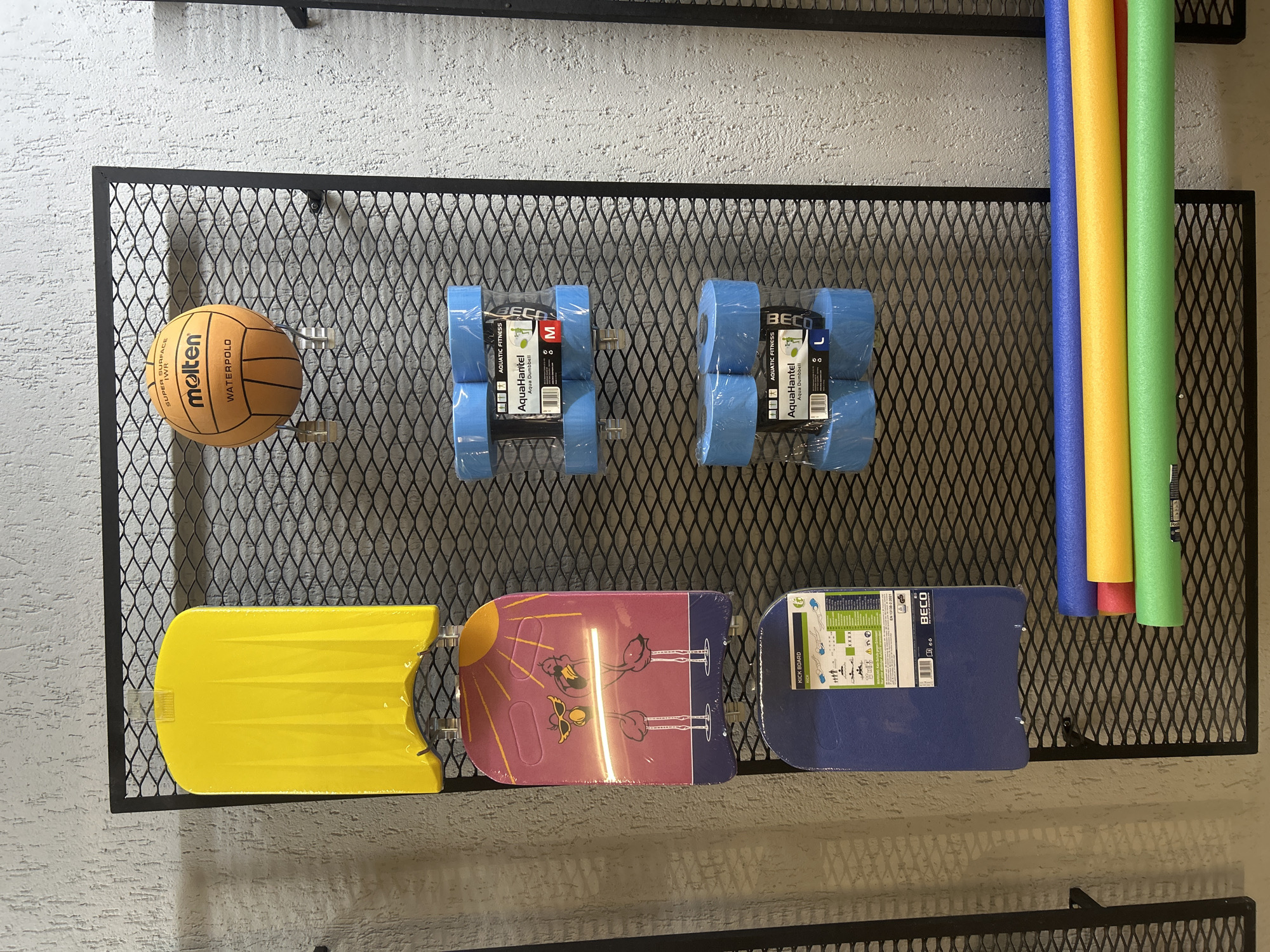 The image shows a black metal rack hanging on a textured, off-white wall. The rack holds various water sports and fitness items. From left to right, the items are: a brown Molten water polo ball, two pairs of blue Beco aquatic dumbbells (one labeled "M" and the other "L"), a yellow kickboard, a pink kickboard with a flamingo design, a blue Beco kickboard, and four pool noodles in blue, yellow, red, and green. The items appear to be for sale or storage. The location is not apparent from the image.