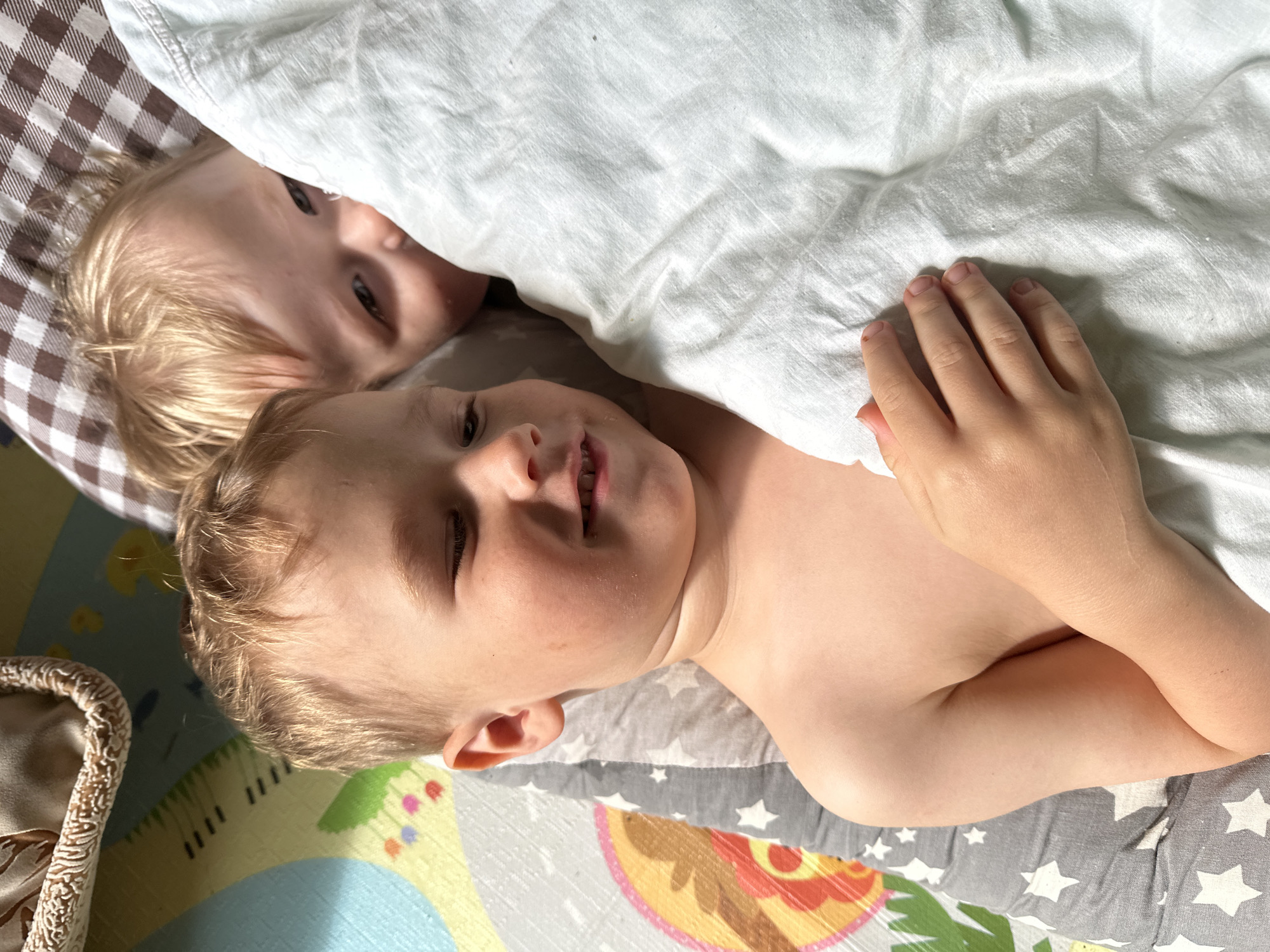 Two children are lying together under a white blanket, partially visible, on a play mat with colorful geometric designs, in what appears to be an indoor setting. One child is more exposed, revealing fair skin and blonde hair. The other child is partially hidden under the blanket, with only a face visible. The background includes a brown and white checkered surface and a textured container. The location and city are not ascertainable from the image.