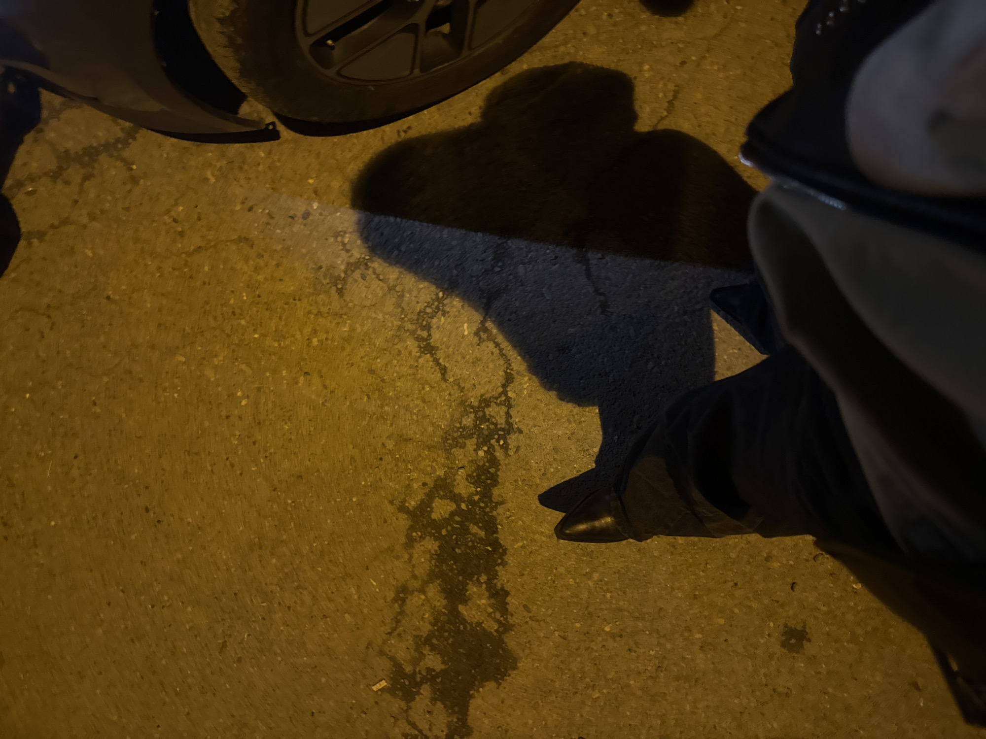 The image presents a low-angle view of a nighttime scene on a paved surface, likely asphalt or concrete, illuminated by an artificial light source that casts prominent shadows. The ground in the foreground has a warm, yellowish hue, and a distinct wet patch with an irregular shape is visible near the center-left.
On the right side of the frame, the lower part of a person's leg and a black shoe or boot are visible. Above the shoe, a hand appears to be holding a smartphone or similar device, partially obscured. A large, dark shadow resembling a person's upper body and head is cast across the central portion of the ground, stretching towards the upper left.
In the upper-left quadrant of the image, parts of a vehicle are visible, including a dark tire with a multi-spoke wheel and a dark fender. The strong contrast between the brightly lit ground and the deep shadows, along with the overall dark background, indicates that the photograph was taken at night. No discernible text is visible in the frame. The scene suggests an urban setting, consistent with its location context in Tbilisi, Georgia.