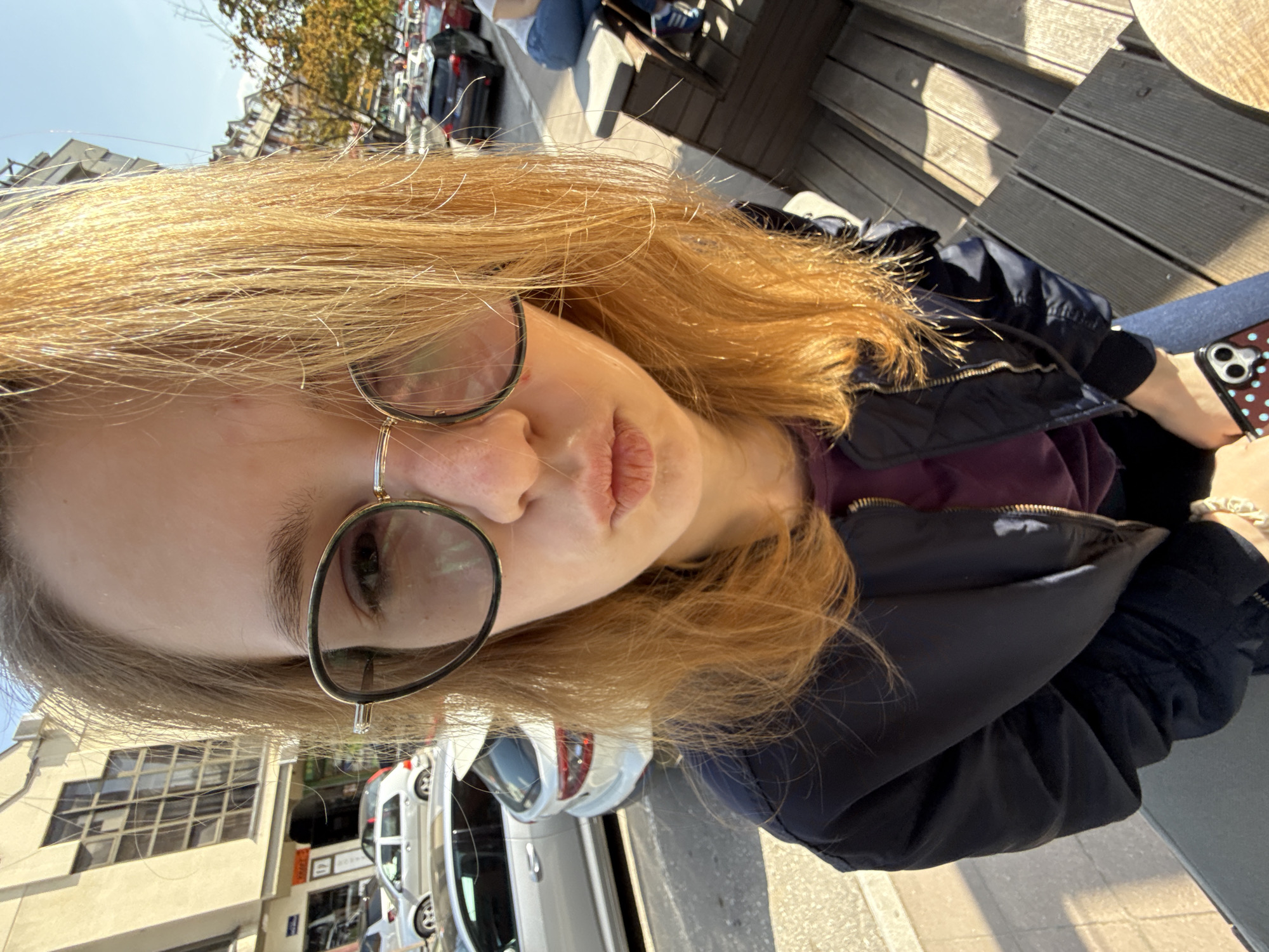 A young woman with long, blonde hair and round wire-frame glasses is captured in a close-up, slightly rotated portrait, seemingly outdoors in Kragujevac, Serbia. She wears a dark blue or black jacket over a purple top. Her lips are slightly pursed, and her expression is neutral as she looks towards the camera. Her right hand is visible, holding a smartphone with a dark, polka-dotted case.
The setting appears to be a sunny day, with bright sunlight illuminating her hair and casting shadows, indicating a clear sky overhead. Behind her, to the right, are dark wooden planks, possibly part of an outdoor seating area or terrace. In the background, to her left, are various buildings, including a light-colored structure with multiple windows. A street scene is also visible, featuring several parked cars (white, silver, and dark vehicles) suggesting an urban environment. There are no clearly legible texts or signs in the image crops.