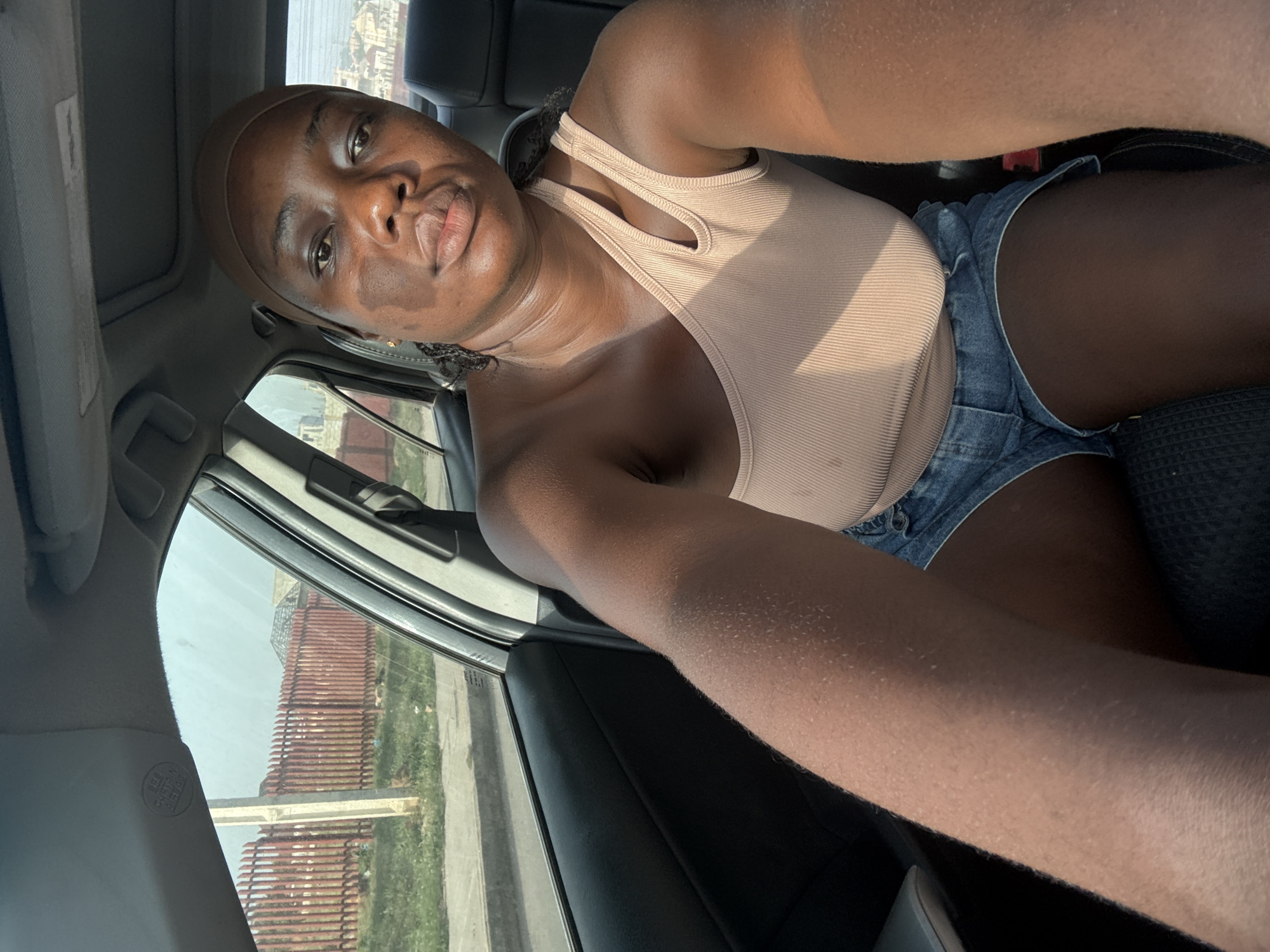 A dark-skinned person takes a selfie from the passenger seat of a car, angled upwards. The person wears a light beige or nude-colored ribbed tank top with a distinctive cutout design near the collarbone and blue denim shorts. They have a matching light brown headwrap or cap and are looking directly at the camera with a slightly pouted expression. Bright sunlight casts highlights and shadows across their skin and clothing.
The car's interior is visible, including a dark seat, a light-colored roof liner, and parts of the window frame. A safety warning label or indicator, showing "SRS AIRBAG" in both English and Japanese characters, is visible on the pillar near the windshield.
Through the car window, an outdoor scene is visible. There's a prominent, tall reddish-brown structure, possibly a building or a fence, characterized by numerous horizontal slats or bricks. Below it, some green grass or vegetation and what appears to be a road or paved area are present. The strong sunlight suggests it is daytime. The setting for this image is Iddo Tudunwada, Nigeria.