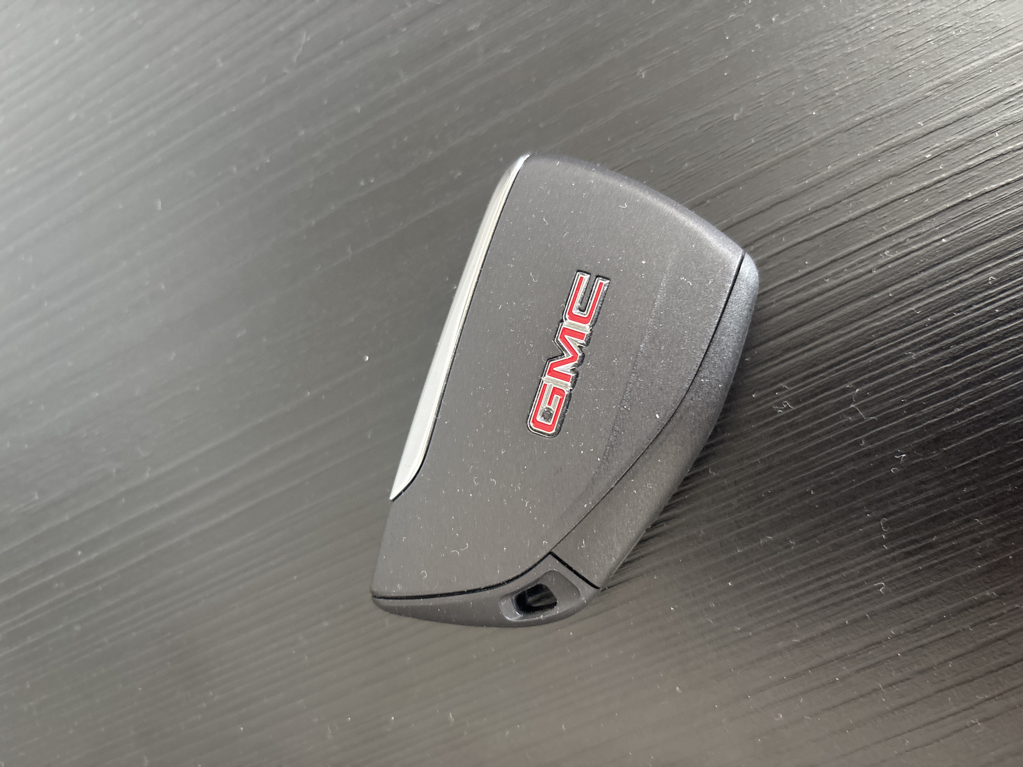 A dark gray, textured car key fob with a red and silver GMC logo is resting on a dark, horizontally lined surface. The key fob is angled, with its rounded end on the left and its flatter end on the right. A small, dark gray loop is visible at the rounded end. The surface it is on appears to be wood or a similar material with distinct vertical grain patterns. The lighting is overhead and casts soft shadows. The overall scene is clean, with no other objects or people present. The location is not discernible from the image; it could be anywhere.