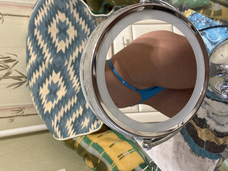 A magnified image captured through a circular mirror reveals a person's buttocks clad in a blue thong. The mirror's chrome frame is visible. The background is a tiled wall. The surroundings include a cushion with a blue and beige geometric pattern and a towel with various colors and patterns. A small faucet is seen on the right side of the mirror.