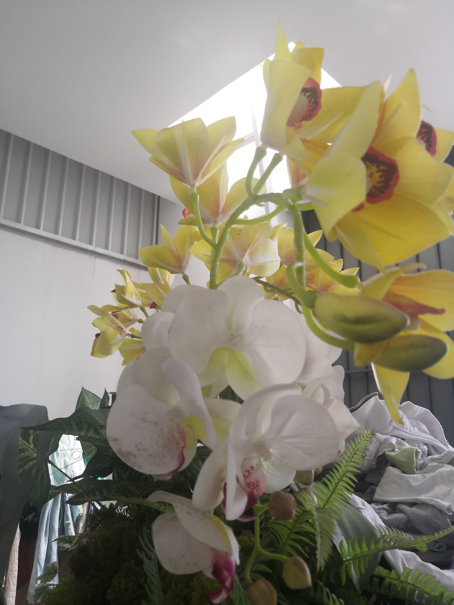 A low-angle, indoor shot showcases a bouquet of artificial orchids, predominantly yellow and white, with some buds. The flowers are arranged in a cluster and occupy the majority of the frame. In the background, a slatted grey wall and a bright, rectangular light source suggest an interior space. To the right, a pile of crumpled white and grey clothing is visible. The scene is likely located in a residential or storage area in Bac Nha Trang Ward, Vietnam, given the context.