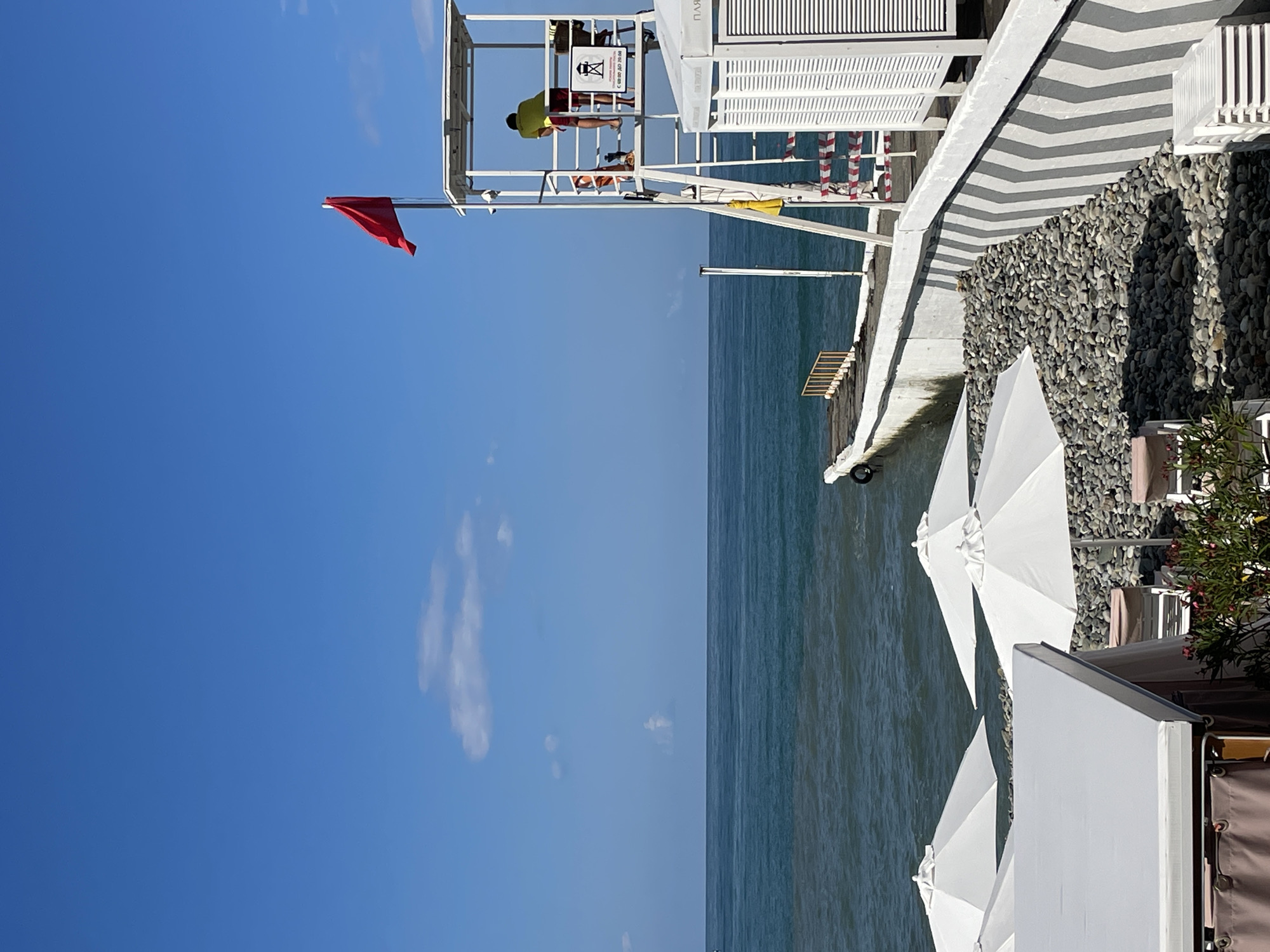 The image depicts a seaside location, likely a beach, with a lifeguard tower prominently featured on the right side of the frame. A red flag is flying from the top of the tower, indicating potentially hazardous conditions or a closure of the swimming area. A person, presumably a lifeguard, is visible on the tower, wearing a yellow shirt and red shorts, and looking out towards the sea. The sea itself is a deep blue-green, with visible waves and a clear horizon. In the foreground, several white beach umbrellas are set up, with lounge chairs visible beneath some of them. The beach appears to be covered in grey pebbles. A concrete pier or walkway extends into the sea from the left side of the frame, with a yellow railing. On the right, next to the lifeguard tower, there are white structures with louvred doors, possibly changing rooms or storage facilities. The sky is a vibrant blue with a few scattered white clouds. The scene conveys a sense of a calm, perhaps slightly restricted, day at the beach. Given the elements present, this scene could be located in Sochi, Russia, a popular Black Sea resort city known for its beaches and recreational activities.