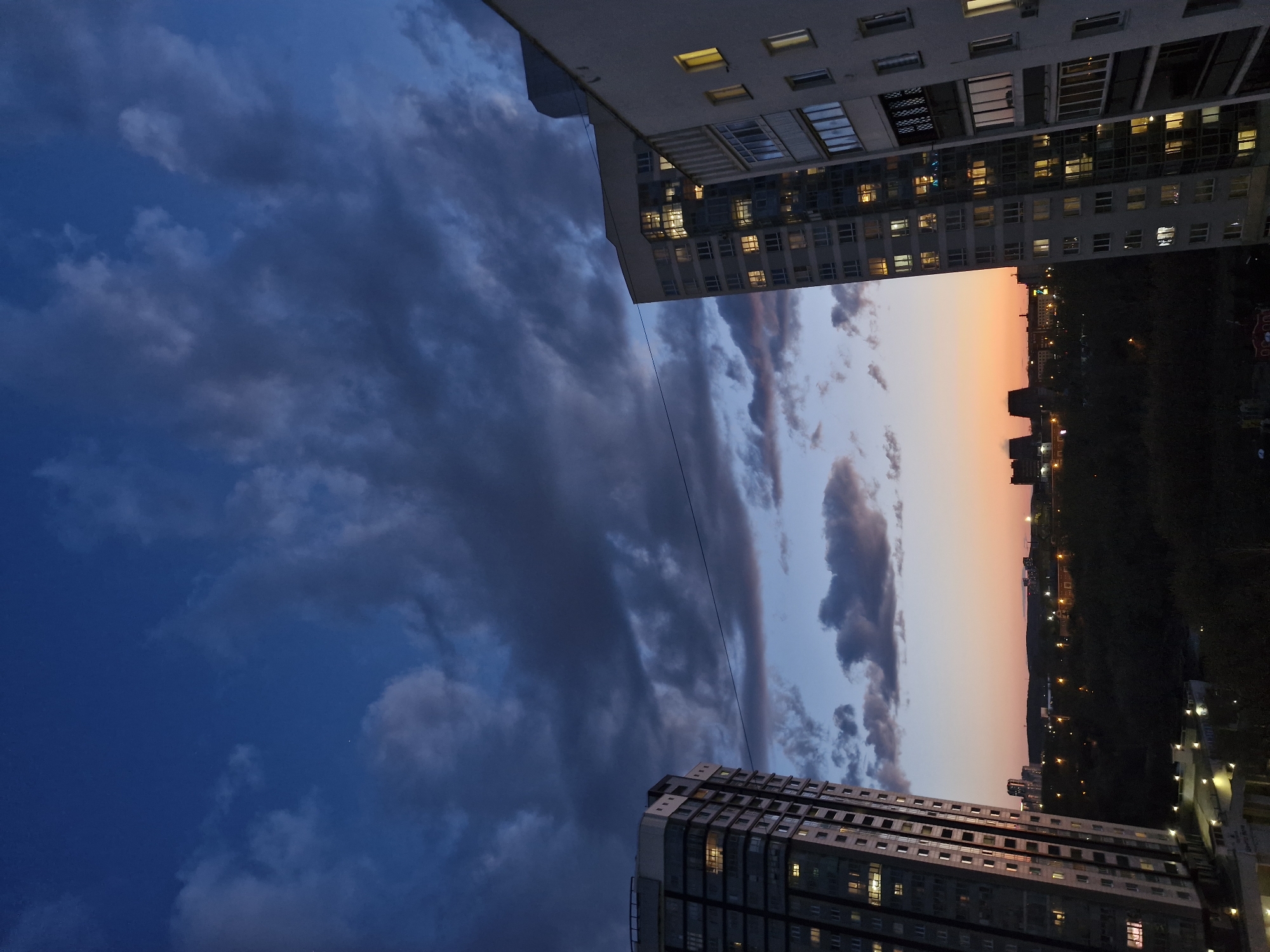 The image captures an urban cityscape at twilight, viewed from above, looking towards the horizon. The photograph is oriented vertically, showing the sky, two buildings, and a distant cityscape.
The sky occupies the upper portion of the image (when oriented correctly). It transitions from a deep, dark blue with heavy, dark grey clouds on the left side, to a softer orange, pink, and light blue gradient on the right side, indicative of a sunset or twilight. Several clouds are visible, some dark and dense, others illuminated by the fading light.
Two large, multi-story buildings frame the view. The top sections of these buildings are visible, one in the upper left corner and another in the lower right. They appear to be modern residential or office blocks, constructed from light-colored concrete or similar materials, featuring numerous windows and balconies. Many of the windows in both buildings glow with warm yellow light, suggesting interior lights are on. A thin, dark wire or cable stretches diagonally across the middle of the sky, running between the two buildings.
On the distant horizon, below the sunset-tinged sky, a silhouette of hills or land is visible, dotted with numerous small, twinkling lights, indicating a distant city or town. The immediate foreground below the horizon is dark, likely comprising trees or undeveloped land.
The time of day is clearly dusk or early night, characterized by the gradient of light in the sky and the illuminated windows and distant city lights. The weather appears cloudy but calm. There are no people or explicit activities taking place, and no visible text. The scene evokes a tranquil, reflective mood of an urban evening. Based on the provided context, the location is Yekaterinburg, Russia.