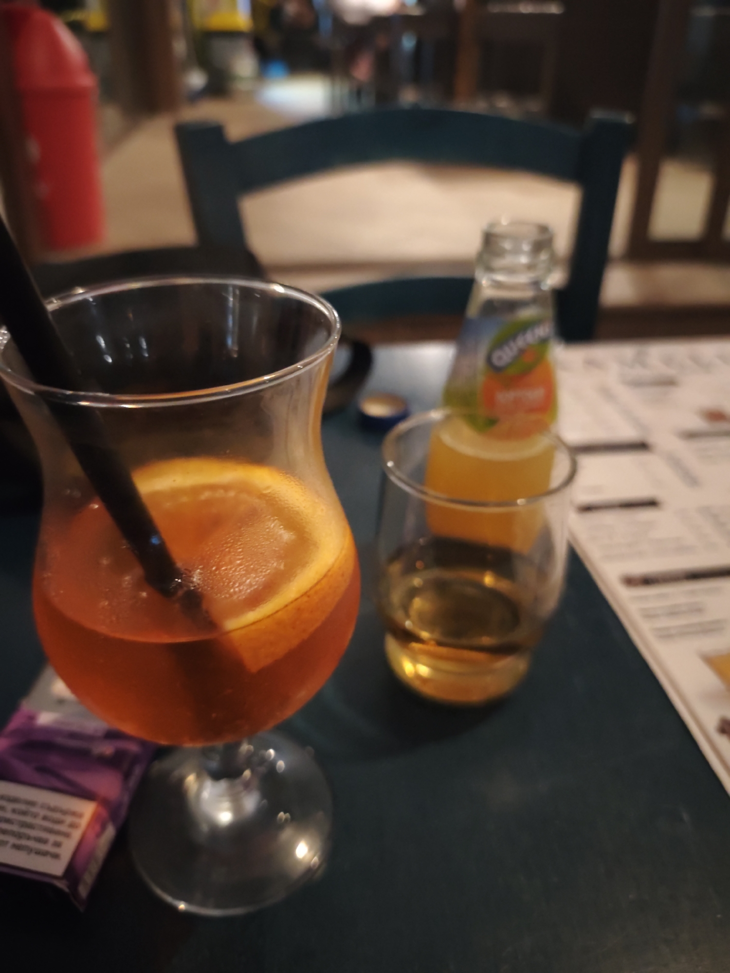 An orange beverage with an orange slice is in a hurricane glass with a black straw. A small glass with a golden liquid, likely alcohol, is to the right of the hurricane glass. Behind these drinks is a small glass bottle with an orange label that reads "QUEEN." A pack of purple cigarettes is to the left of the hurricane glass. The scene appears to be set on a dark blue table, possibly in a bar or restaurant. In the background, blurred chairs and a red object are visible. The image is likely taken in Nesebar, Bulgaria, given the reference to its location in the prompt.