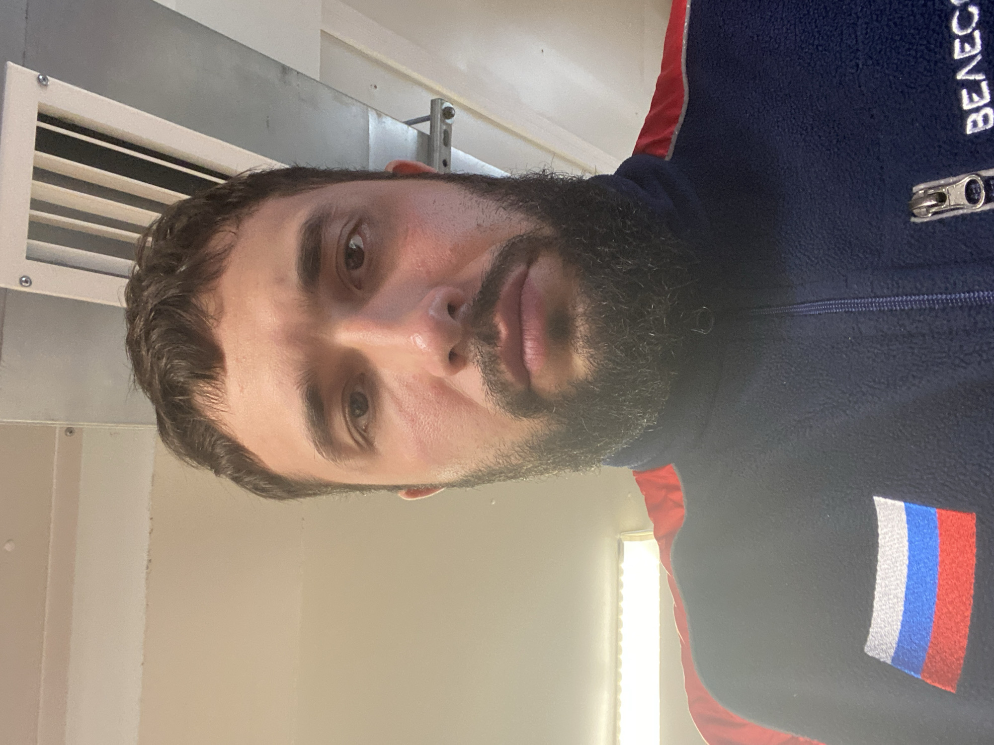 A man with a dark beard and short, dark hair is shown from the chest up in a close-up selfie. He is wearing a dark blue fleece jacket with red accents on the shoulders and a Russian flag patch on the left chest. A zipper is visible on the jacket. The background appears to be indoors with a textured ceiling and a ventilation grate. The lighting is bright, casting some shadows. The scene is static, with no action or interaction depicted. The location is identified as Luzhitsy, Russia.