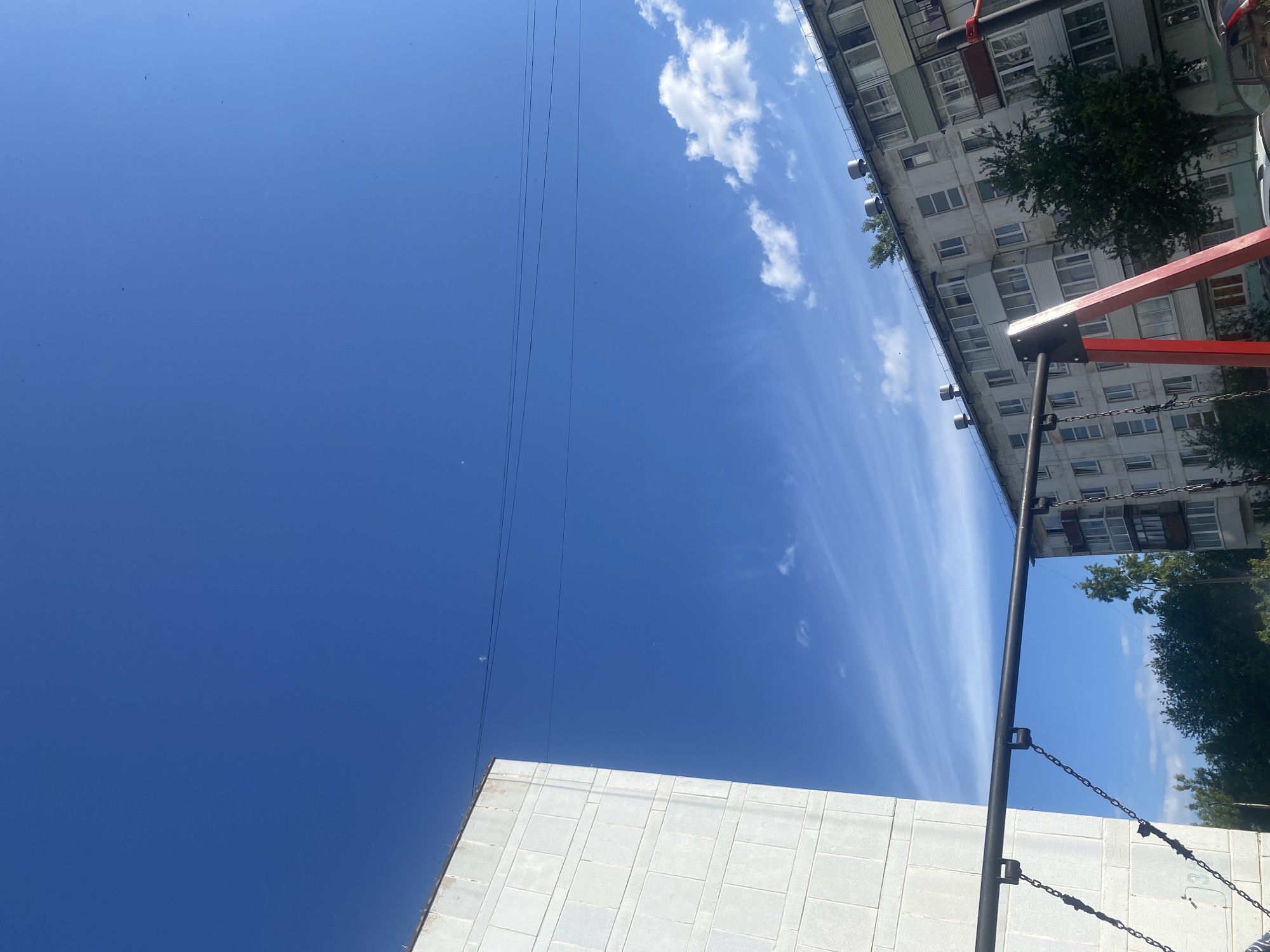 The image depicts an outdoor urban scene in the city of Chaikovsky, Russia, characterized by a predominantly clear, deep blue sky with scattered white cirrus and cumulus clouds. Two distinct multi-story buildings frame the scene. On the left, a tall, light-colored, block-patterned building occupies the vertical left third of the frame. On the right, a grey, weathered apartment building with multiple windows, balconies, and several ventilation units on its roofline extends across the mid-right. Two dark utility lines traverse the upper-middle sky horizontally. In the immediate foreground, a dark metal horizontal bar, suspended by chains, extends across the lower portion of the image. This bar is supported on the right by a bright red A-frame structure, indicative of a swing set or playground equipment. Green foliage and tree tops are visible between and below the buildings, suggesting landscaping or nearby natural areas. No individuals are visible. The overall lighting suggests daylight conditions.