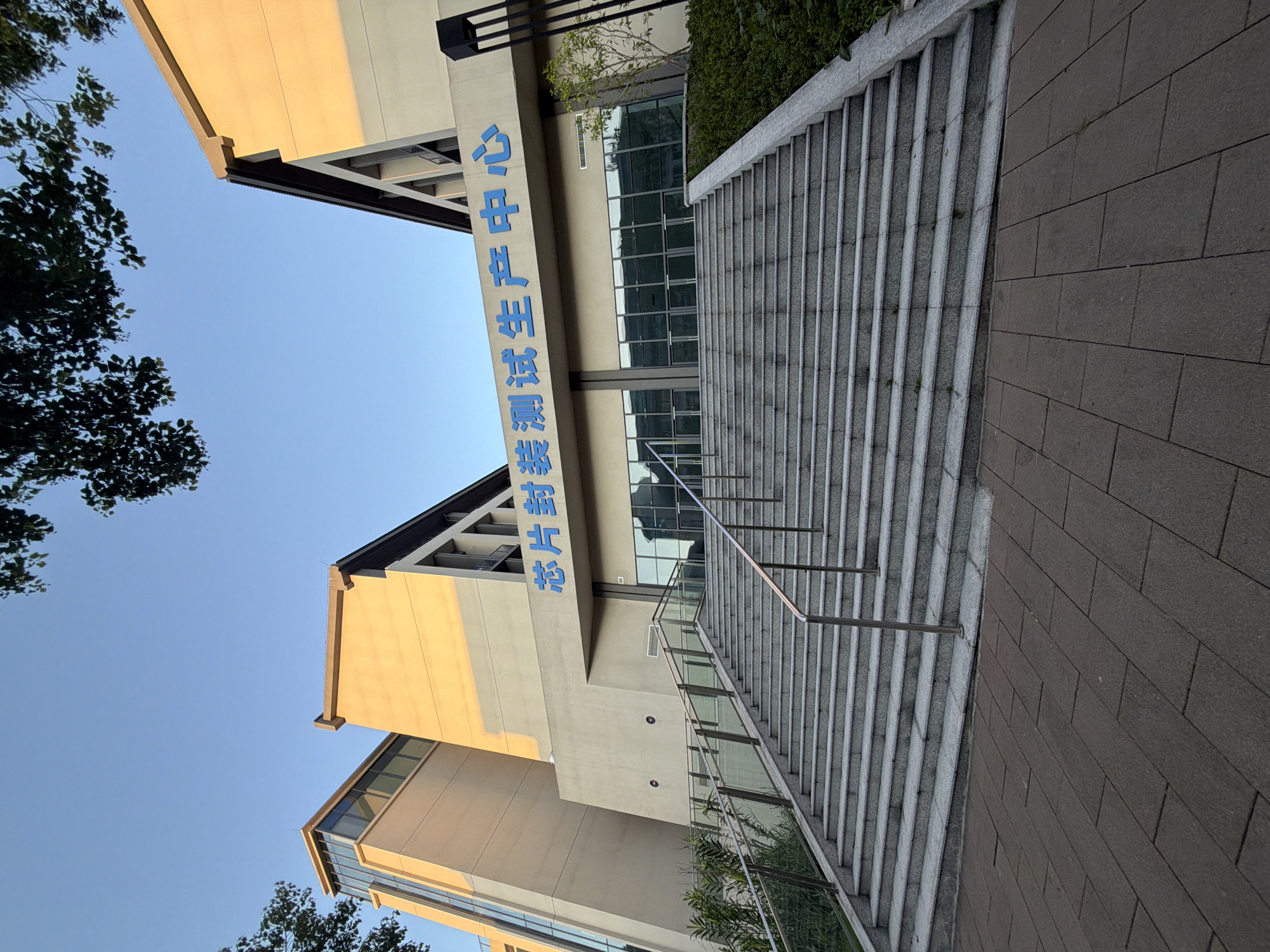 The image shows a modern building with a wide staircase leading to its entrance. The building is light beige with angled, dark-roofed sections. Blue Chinese characters are visible above the entrance, which consists of large glass doors. The staircase is made of grey stone and has metal handrails. A paved walkway is in the foreground. Greenery flanks the staircase and surrounds the building. The sky is clear and blue, and trees with dark leaves are visible at the top of the frame. The building is identified as the "Chip Packaging and Testing Production Center" in Jiangjin, China. There are no people in the image.