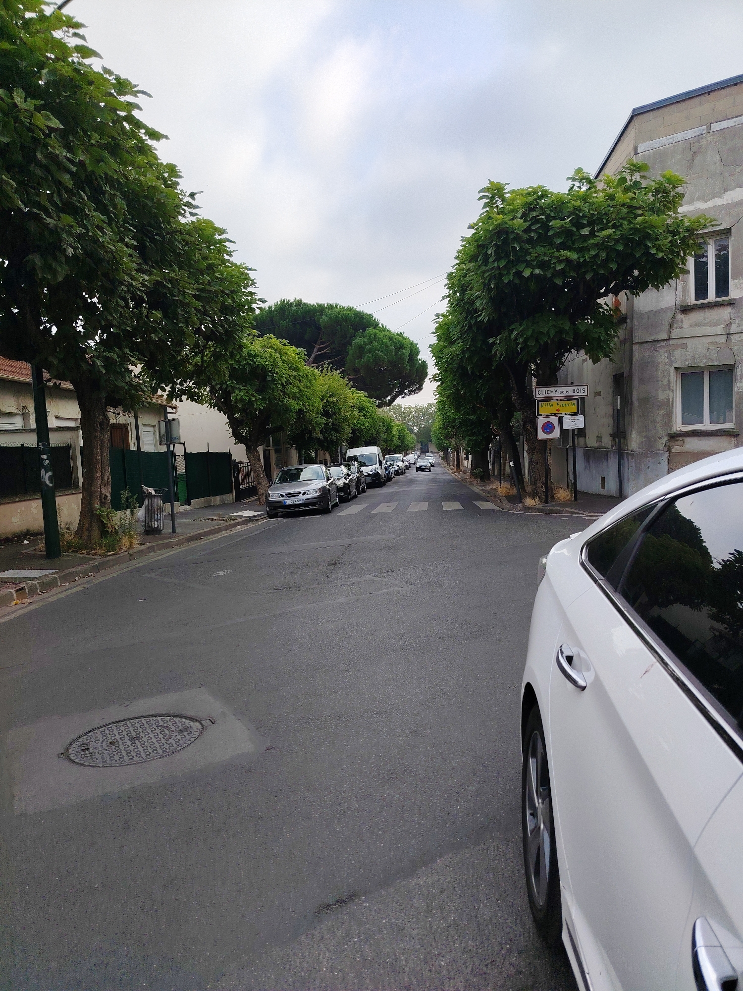 The image depicts a street scene in Clichy-sous-Bois, France, under an overcast sky. In the foreground, the right side of a white car is visible, showing its side door, door handle, and wheel. The car is parked or stopped on the right side of a paved road.

The road itself is lined with trees on both sides. On the left, houses with dark green fences and a manhole cover set into the pavement are visible. On the right, a multi-story building with a cracked concrete facade and windows stands next to the road, partially obscured by trees. A street sign reads "CLICHY-SOUS-BOIS" and another sign indicates "Ville Fleurie."

Several cars are parked along the left side of the street, including a silver sedan with a license plate that appears to read "WL-087-LH". Further down the road, more cars are parked, and the street recedes into the distance, lined with trees. The overall impression is a quiet, residential street in a suburban setting.