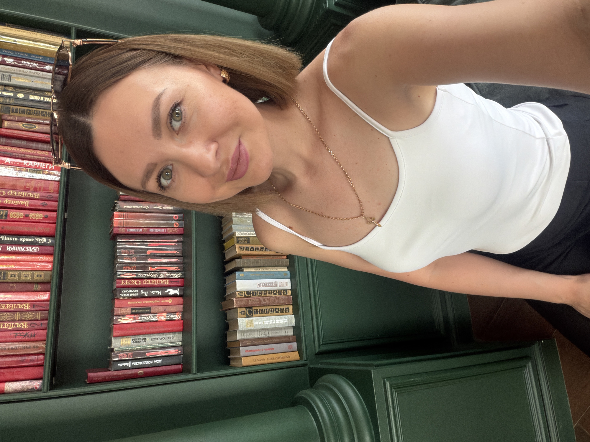 A woman wearing a white tank top and gold necklace is taking a selfie. She is standing next to a green bookshelf filled with books. Sunglasses are perched on top of her head. The bookshelf appears to be part of the interior of a building in Rostov-on-Don, Russia, based on the Russian titles of the books on the shelves.