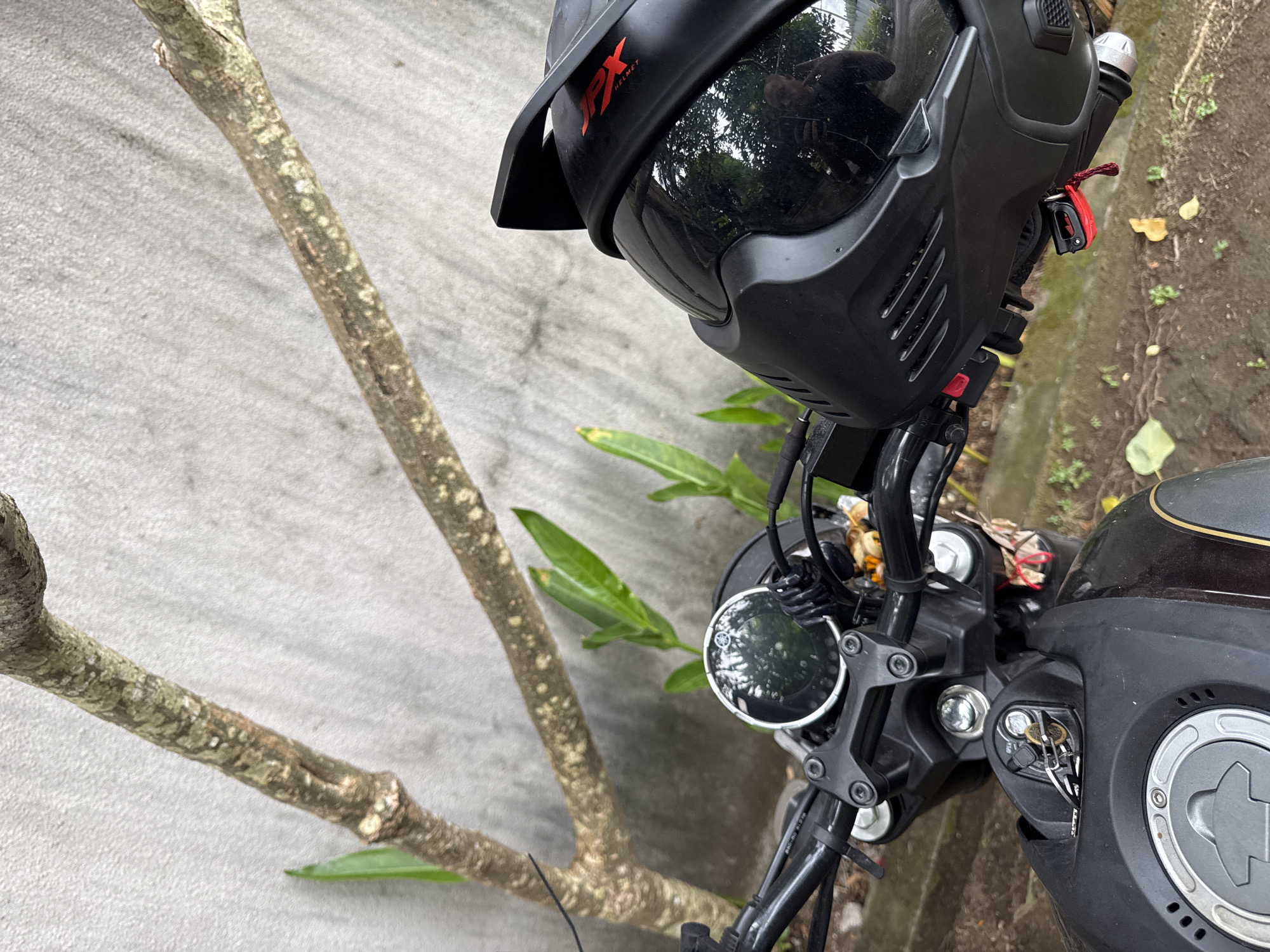 The image shows a close-up, high-angle view of a black motorcycle with a helmet resting on its handlebars. The motorcycle has a round rearview mirror and a digital display. A key is in the ignition. The helmet is black with a red "JPX HELMET" logo. The scene appears to be outdoors, with a grey textured wall in the background and some greenery. The ground is dirt and leaves. The image was captured in Kuta, Indonesia.