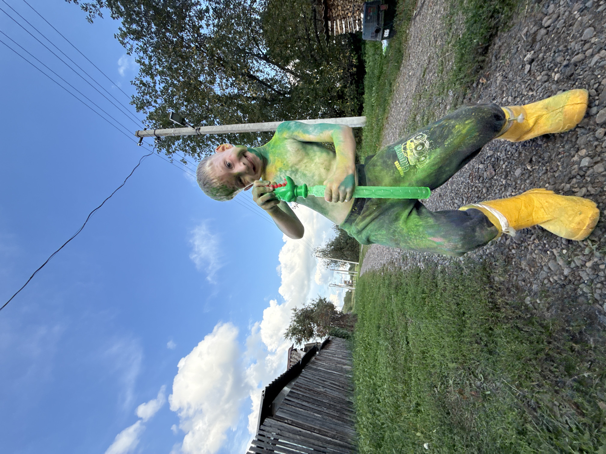A young Caucasian boy, approximately 8 years old, is standing on a gravel road in Nizhnyaya Chermoda, Russia. He is partially undressed, with green and yellow powder paint covering his torso and legs. He is wearing green pants with a monster truck graphic and bright yellow rubber boots. In his hands, he holds a green plastic bubble gun. The boy has a playful expression and is looking at the camera.
The background features a wooden fence on the left side of the image, with a grassy field bordering the fence. Beyond the fence, trees and some rural structures are visible. The road continues into the distance, flanked by vegetation. A utility pole with power lines stands to the right of the boy. The sky is blue with scattered white clouds. The overall impression is of an outdoor, informal setting, possibly a village or rural area in Russia.