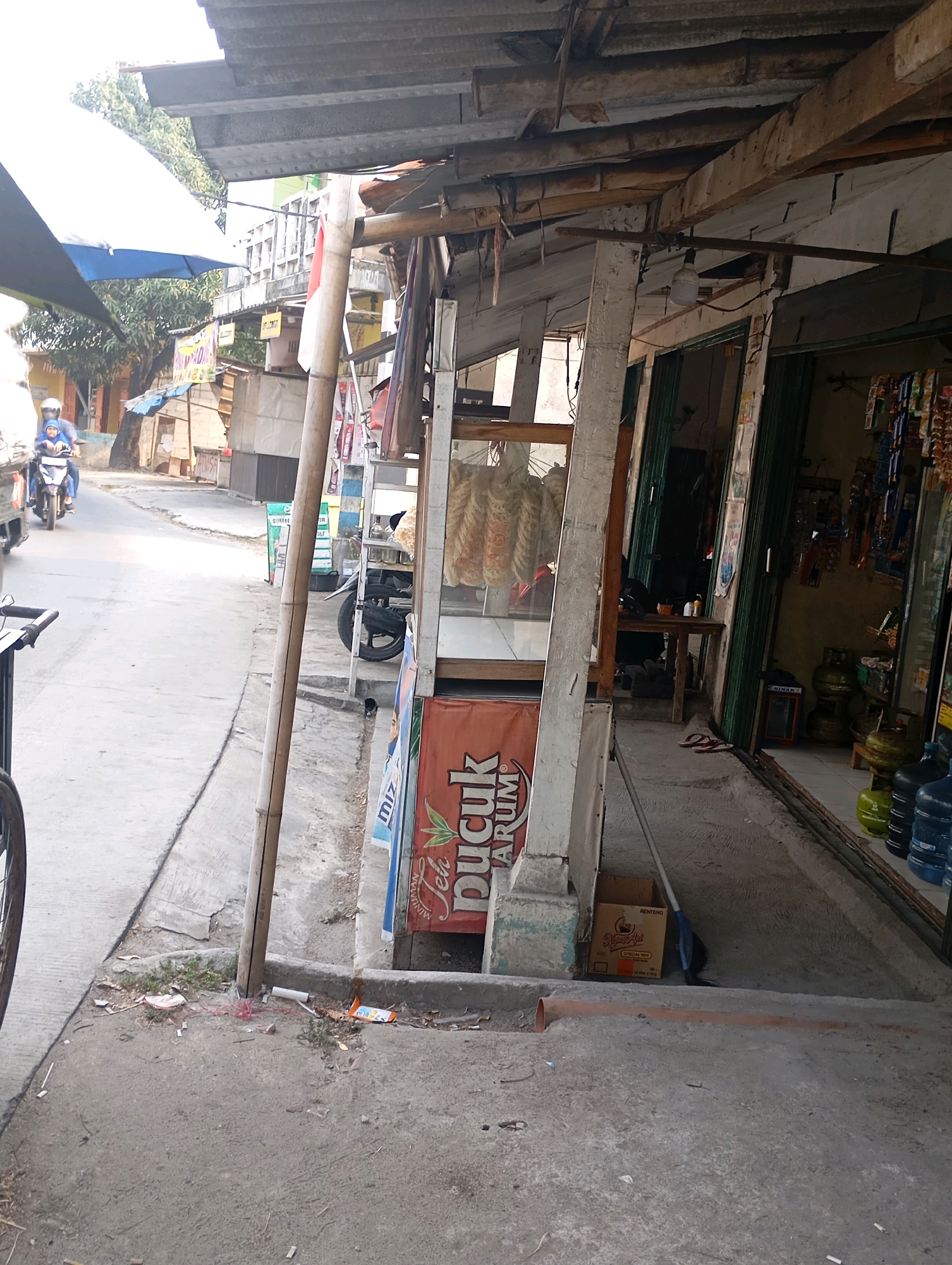 The image captures a street scene in Babelan, Indonesia, with a focus on a roadside stall. A person on a motorcycle, wearing a blue shirt and a helmet, is seen riding down the street. The stall is constructed with wooden beams and corrugated metal roofing, typical of many informal businesses in the region. In front of the stall, a display case holds various items, possibly food products, with one prominent sign featuring text that appears to be in Indonesian. Next to the stall, several gas cylinders and large water bottles are stacked, suggesting they are for sale. A broom leans against a cardboard box. The street itself is paved, with a concrete sidewalk bordering the stall. The overall environment suggests a bustling, everyday commercial area.