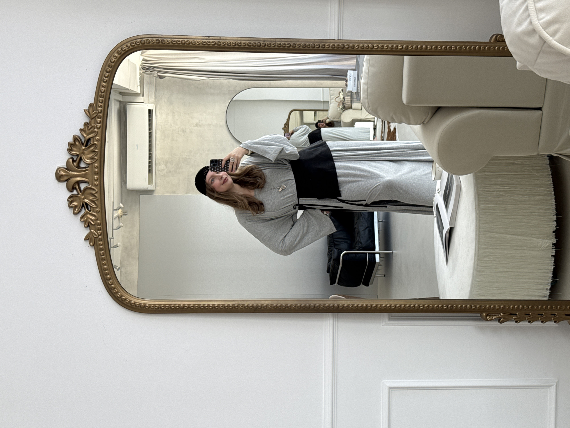 A person is taking a mirror selfie in a room that appears to be a home or a studio. The person is wearing a grey dress with wide sleeves and a black belt cinched at the waist, as well as a black beret. They are holding a smartphone in their right hand. In the reflection of the mirror, we can see more of the room, including a black leather sofa, a white rounded ottoman with fringe, and a cream-colored sofa. There is also a smaller, arched mirror in the background reflecting another person, and a window with sheer curtains. The overall aesthetic of the room is modern and minimalist, with a focus on neutral colors.
The image does not contain any specific visual cues that would allow for the determination of its location within the city of Izhevsk, Russia.