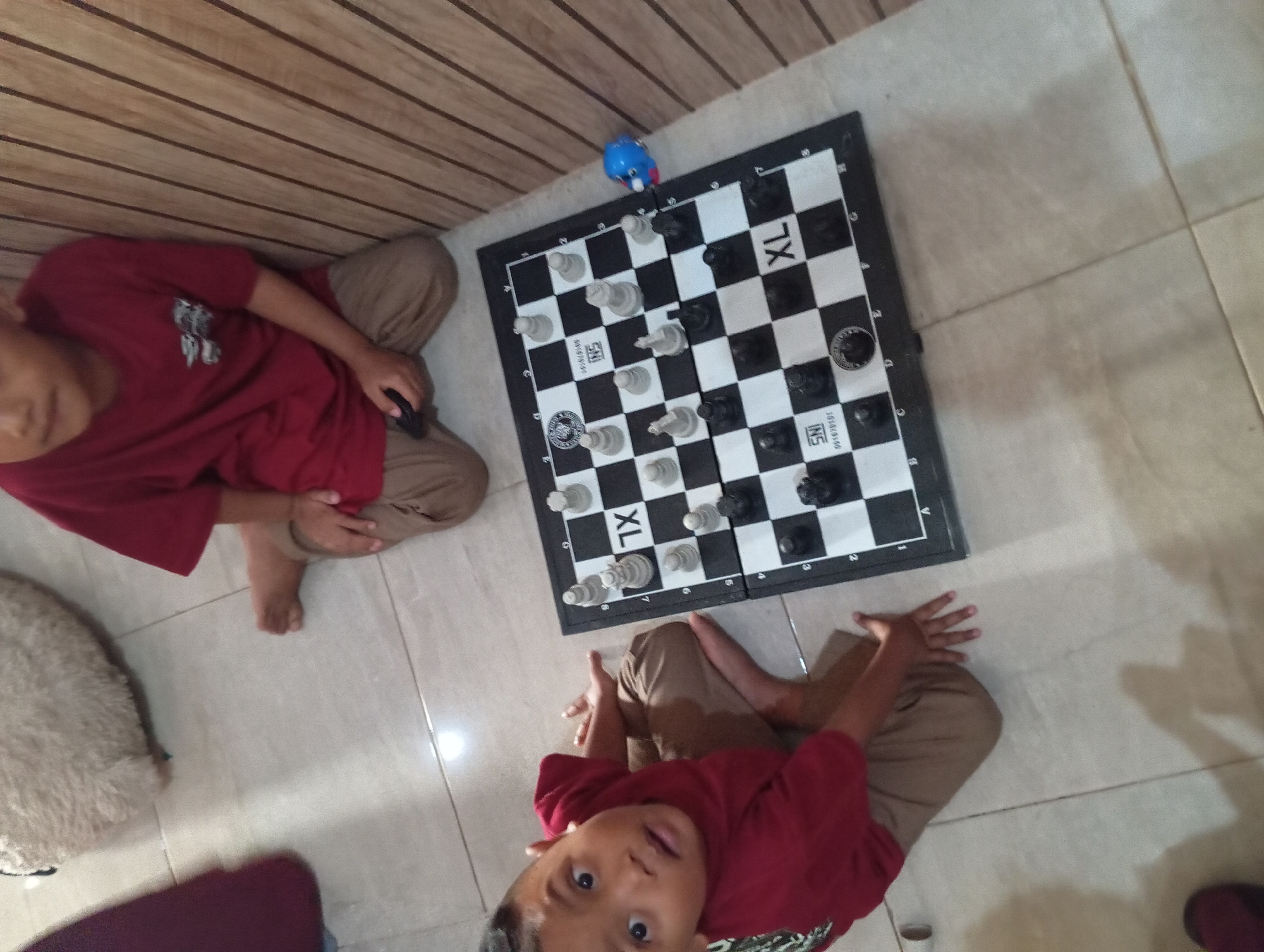 In Padurungan, Indonesia, two young boys are playing chess on the floor. One boy, wearing a red shirt and brown pants, is seated and looking at the camera with a surprised expression. The other boy, also in a red shirt and brown pants, is seated slightly further back, facing the chessboard. The chessboard is set up on a black and white mat with the letters "XL" printed on it. Chess pieces, both white and black, are arranged on the board. A small blue toy is positioned on the edge of the chessboard. The environment appears to be indoors, with a wooden-paneled wall in the background and a tiled floor.