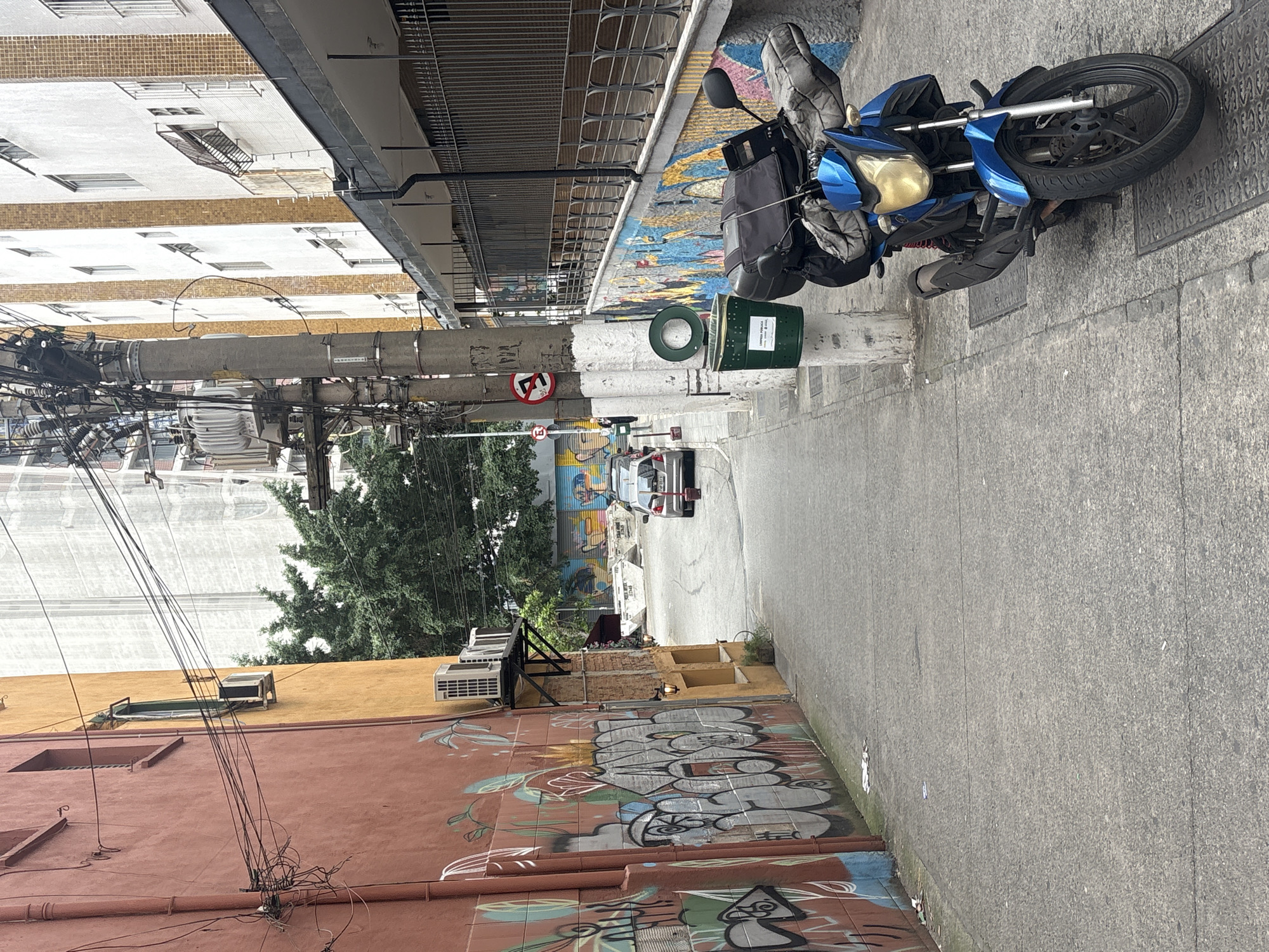 This is a photograph taken in an alleyway in São Paulo, Brazil, specifically in the Vila Madalena neighborhood. The scene is urban and somewhat gritty.
On the left, a building with a rust-colored facade is covered in graffiti. To the right of this, a yellow building has an air conditioning unit mounted outside. The alley itself is paved with concrete.
Dominating the right foreground is a blue motorcycle, parked on the side of the alley. It has a large black storage box mounted on the back and grey covers over its handlebars. A green trash bin is affixed to a utility pole next to the motorcycle.
Further down the alley, a silver car is visible, parked. The alley is lined with buildings on both sides, some with windows and balconies, and a tall utility pole with numerous wires and a transformer is prominent in the mid-ground. The walls of the buildings along the alley are adorned with more colorful murals and graffiti. Trees are visible in the background, suggesting a somewhat dense urban environment. The sky is overcast.