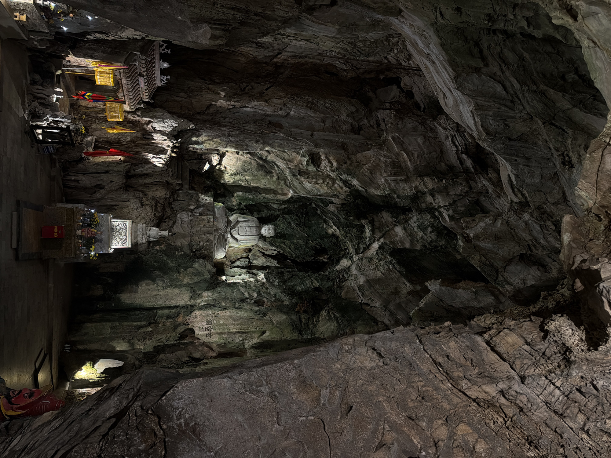 The image shows a section of a cave temple in Đà Nẵng, Vietnam. A prominent white Buddha statue is situated in the center, illuminated by a spotlight. To the left of the statue, there is an altar with offerings and what appears to be a temple structure with golden and red decorations. The cave walls are dark and rocky, with textured surfaces and varying shades of gray and brown. The overall lighting is low, emphasizing certain areas with artificial light while leaving others in shadow.