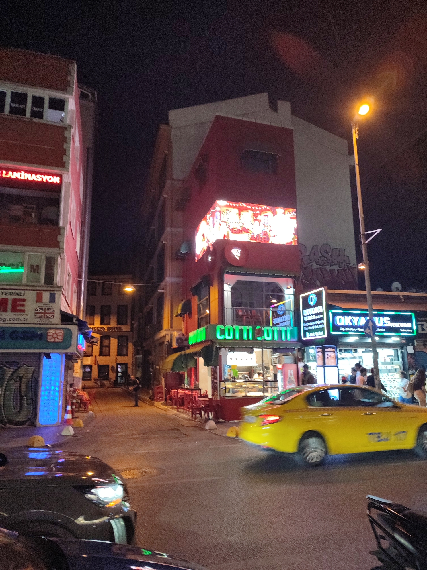 The image depicts a street scene at night in Kadıköy, Turkey, likely a commercial area with shops and restaurants. A bright yellow taxi is captured in motion, its headlights illuminated, moving down the paved street. Several businesses line the street, displaying prominent neon signs. One sign reads "COTTII COTTI" in green lettering, indicating a food establishment. Other businesses feature signs in Turkish, such as "LAMINASYON" and "OKYAYUS TELEKOM".
People are visible in the scene, some standing outside shops, while others are seated at outdoor tables, suggesting a vibrant nightlife. The architecture appears to be a mix of modern and older buildings, common in urban environments. Streetlights and the illuminated signs cast a warm glow on the surroundings, creating a typical urban night ambiance. The street is relatively narrow, with buildings on both sides. The overall impression is of a busy commercial street in a Turkish city at night.