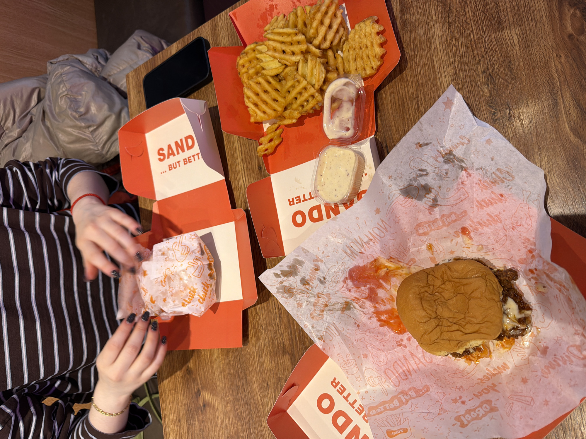 A tabletop view features an array of fast food items: two burgers, waffle fries, and accompanying sauces, suggesting a meal setting. The food is partially unpacked from red and white containers labeled "Sando...But Better", placed on a wooden surface. A person with a brown and white striped shirt is visible, hands raised near one of the burgers. Adjacent to the food are a dark cell phone and a shiny coat. The location is indicated by the writing on the containers, identifying it as "Sando," a fast-food establishment. The images do not indicate what city in Bulgaria this is located in.