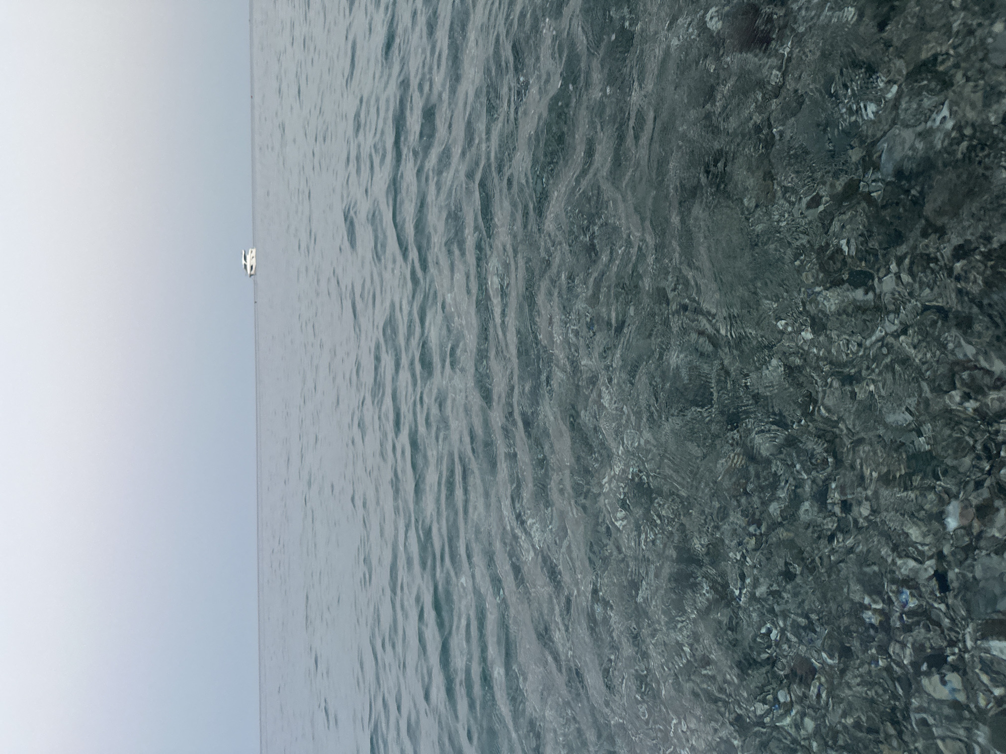 The image depicts a serene seascape with a clear horizon under an overcast sky. In the distance, a white motor yacht is visible, positioned on the tranquil water. The foreground reveals the seabed, composed of various stones and pebbles, visible through the transparent, shallow water. The overall impression is one of calm and clarity, typical of a quiet coastal area. The location is identified as Kemer, Turkey, suggesting a scene from its coastline.