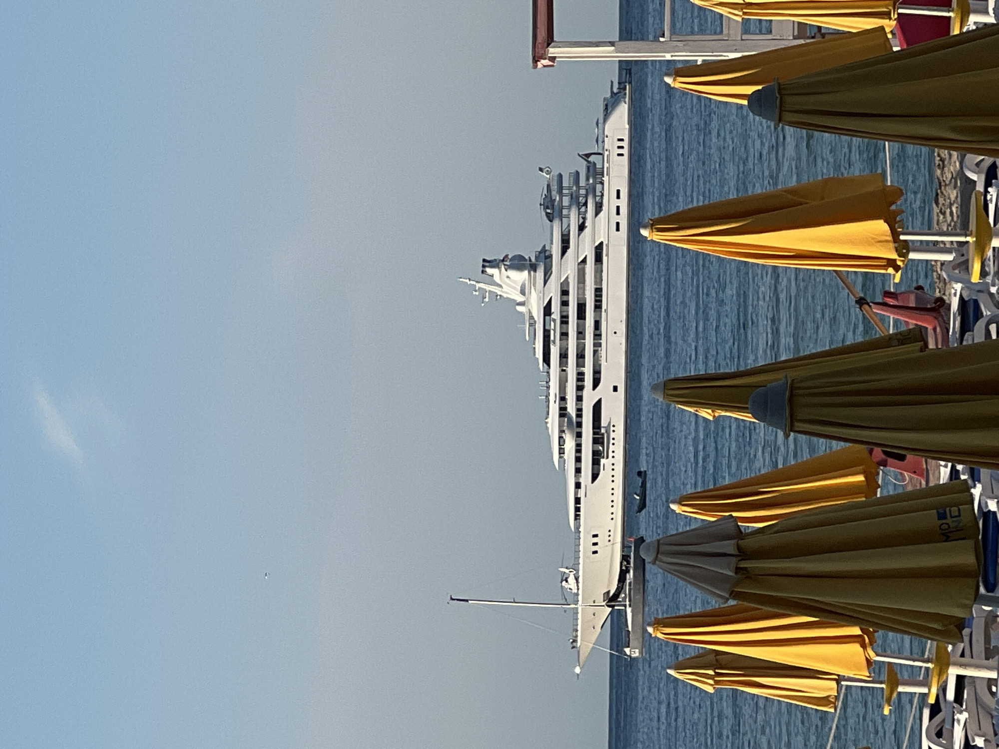 A large, multi-deck white yacht with a helicopter on its deck is anchored offshore under a clear blue sky. In the foreground, several yellow beach umbrellas are partially closed, some showing logos. The sea is calm and blue, with a few smaller boats visible near the yacht. The scene is reminiscent of a sunny day at a beach resort, potentially in a coastal city like Palermo, Italy.
