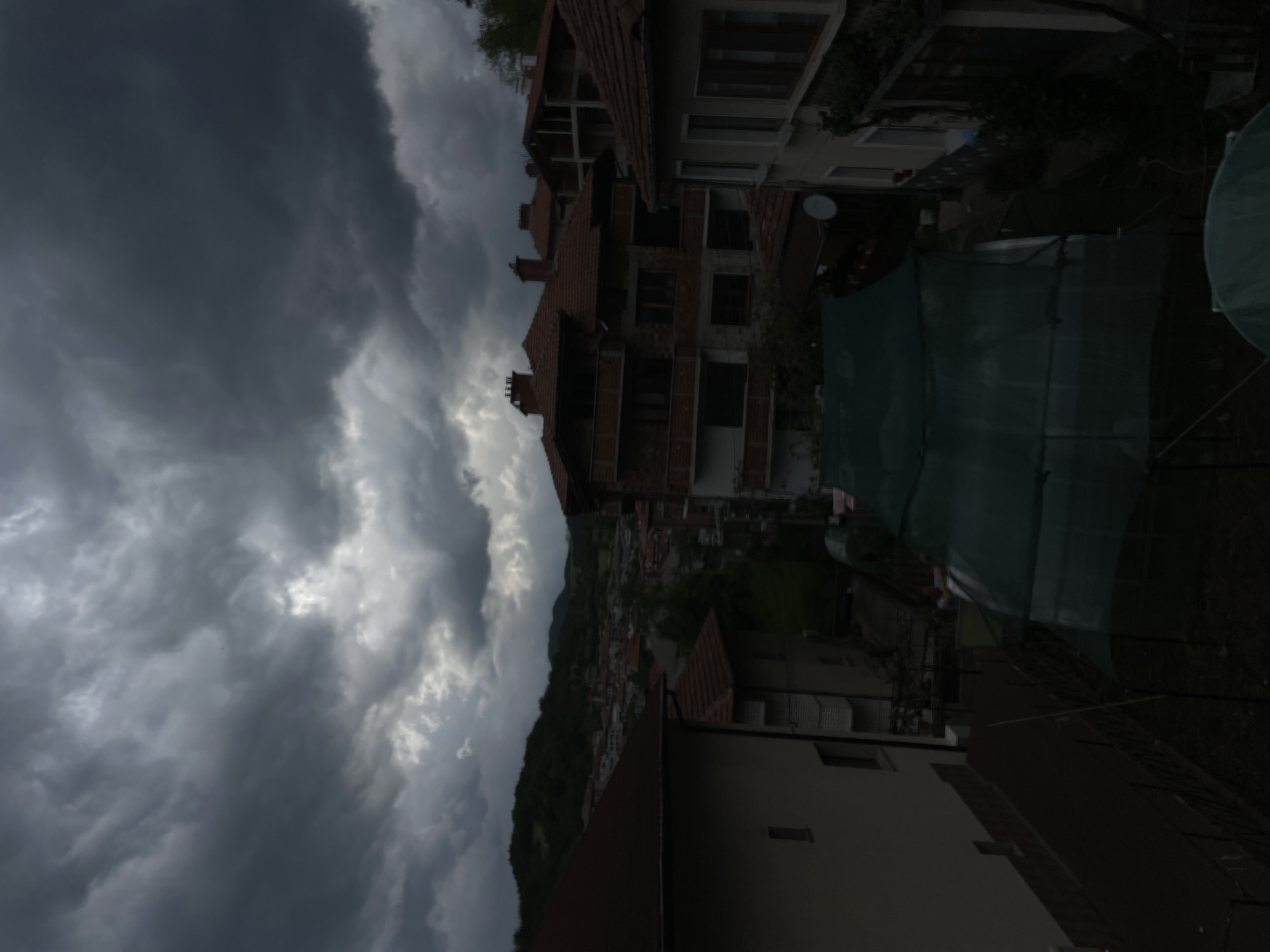 The image depicts a cityscape under a dramatic, stormy sky. The city, likely Zlatograd, Bulgaria, is nestled against a hillside covered with green vegetation. The architecture is a mix of low-rise buildings with red-tiled roofs, characteristic of the region. Dominating the foreground is a multi-story building with exposed brickwork, suggesting an unfinished or ongoing construction project. A satellite dish is visible, mounted on the building. A dark, partially obscured structure, possibly a greenhouse or covered area, sits in the right middle-ground. The sky is filled with dark, swirling storm clouds, creating a somber and ominous atmosphere. The overall scene is still and quiet, with no visible human activity or interaction.