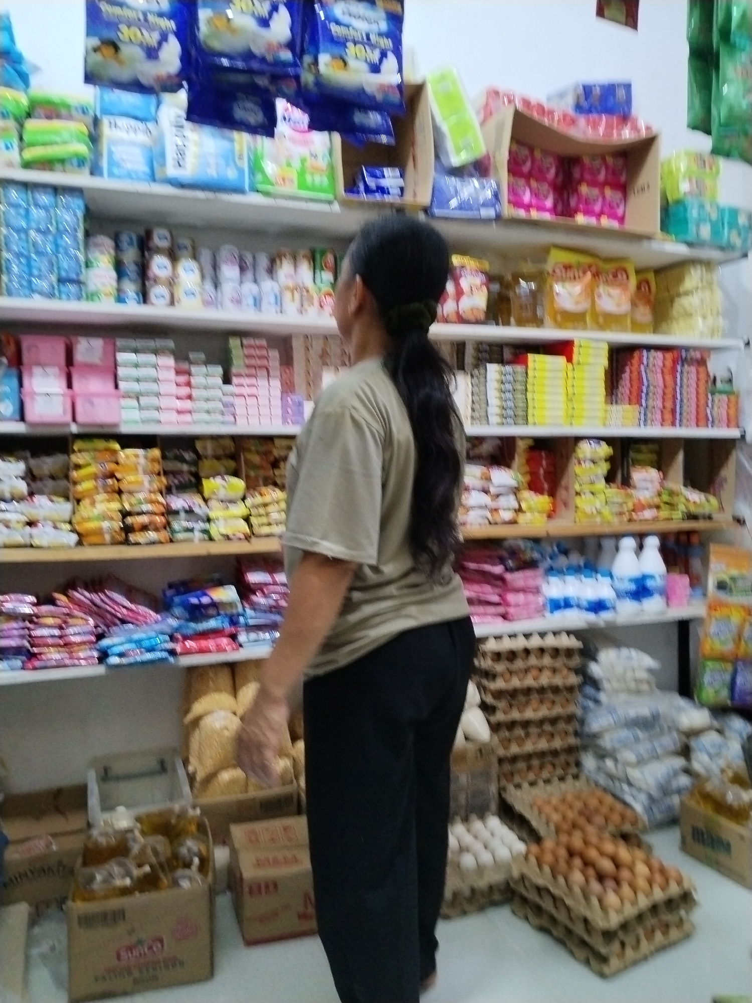 The image depicts the interior of a small grocery store, likely a warung or minimarket, in Sawit Seberang, Indonesia. A woman with long black hair pulled back is standing in the foreground, facing away from the camera, her right hand extended towards a shelf. She is wearing a tan t-shirt and dark trousers. The store is densely packed with shelves filled with various products. On the shelves, one can see a variety of packaged goods, including what appear to be cleaning supplies, food items, and beverages. Specific items visible include stacks of detergent boxes, bottled drinks, packets of snacks, and some larger items like oil bottles in a cardboard box on the floor. The store appears to be well-stocked, with a significant quantity of goods arranged neatly on the shelves and in piles on the floor. The overall impression is that of a typical Indonesian local store, with a focus on essential household and food items.