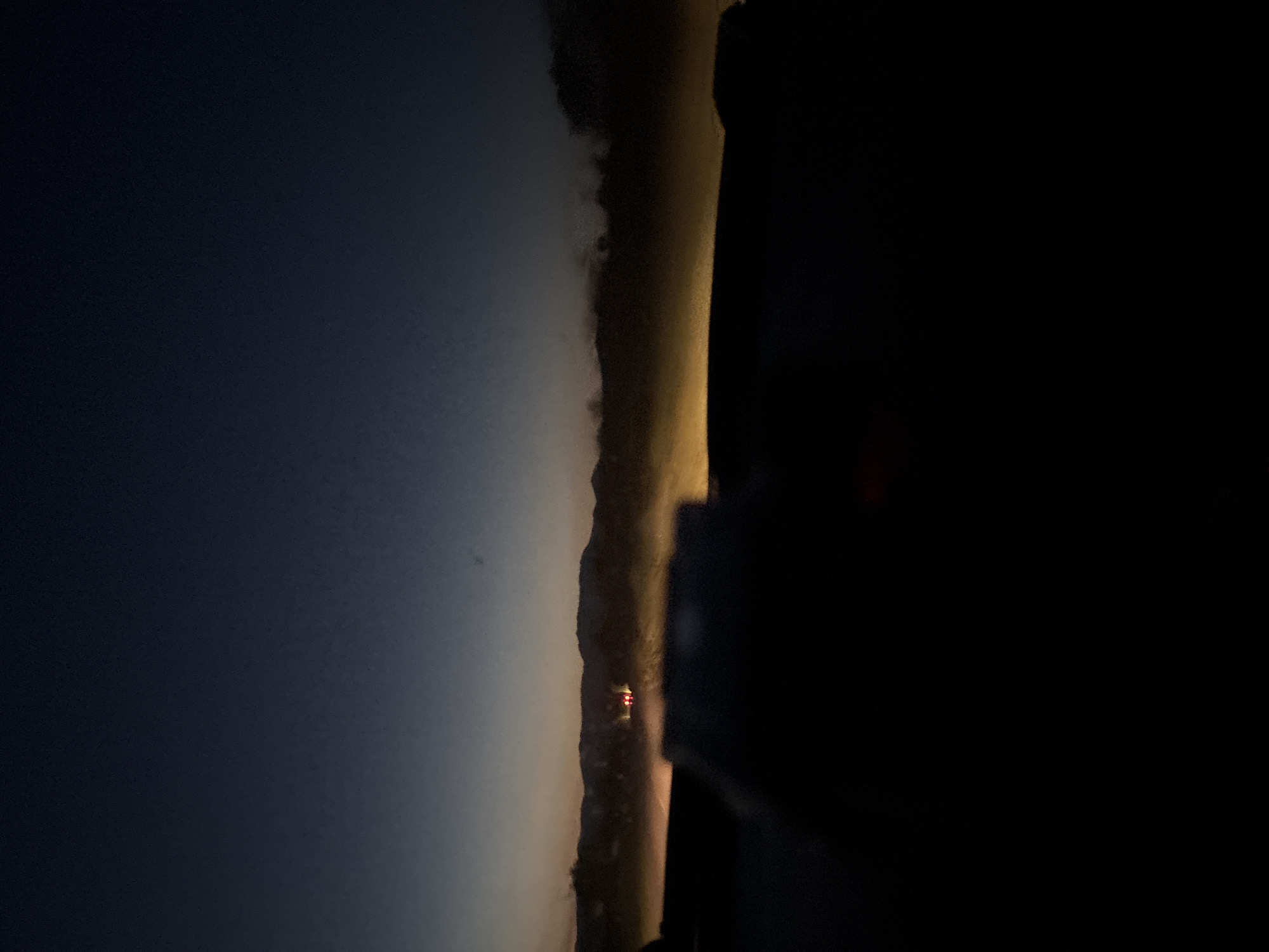 The image depicts a night scene in Novo Zhelezare, Bulgaria. The left side of the image shows a dark blue sky. A lit roadway or path is visible in the center of the image, with a glimpse of red lights potentially from a vehicle or structure. The right side of the image is obscured in darkness.