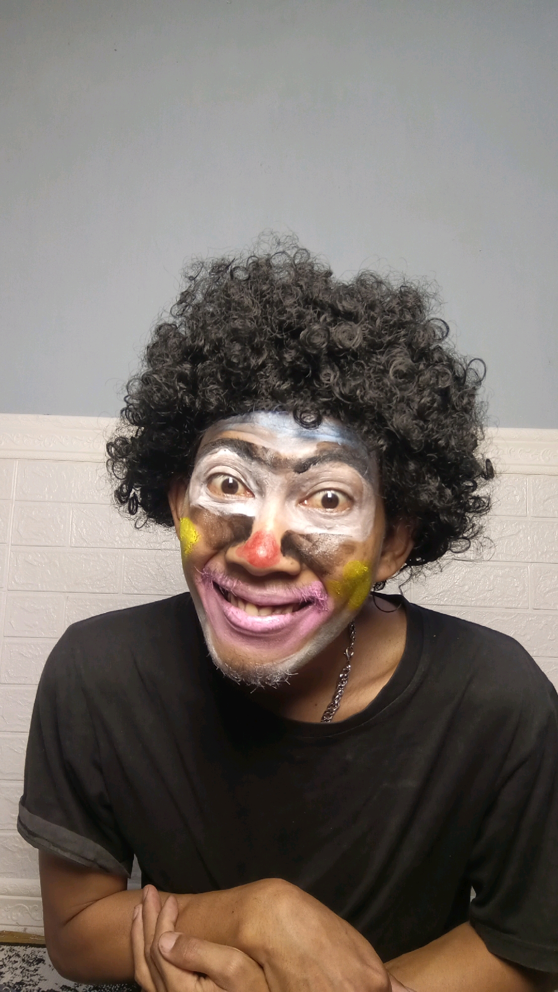 A person with curly black hair and clown makeup is looking directly at the camera in Kebumen, Indonesia. The makeup includes white paint on the forehead and around the eyes, yellow splotches on the cheeks, a red nose, and pink lips. The person is wearing a black t-shirt and a silver chain. They are sitting in front of a light blue wall and a white brick wall. Their hands are clasped in their lap.