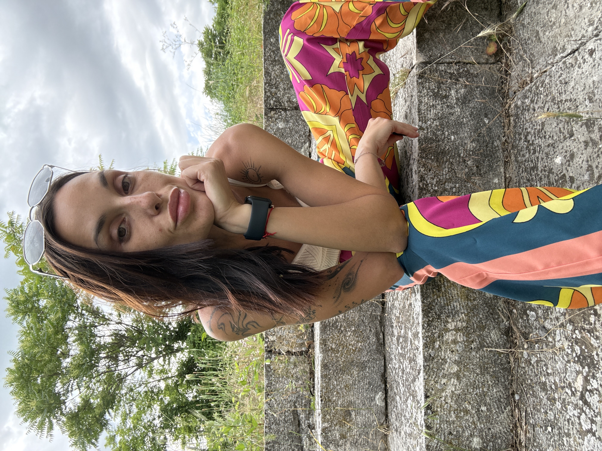 A woman with long, dark hair and sunglasses perched on her head sits on stone steps outdoors. She is wearing a white top and colorful pants with a psychedelic pattern. Her left arm is bent, with her hand resting on her chin, and her right arm is resting on her lap. The background shows greenery and a cloudy sky. The stone steps are weathered and overgrown with grass. The image appears to be taken on a warm, overcast day.